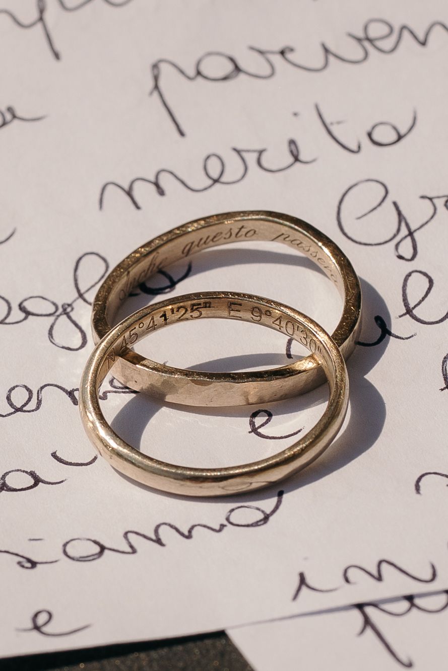 Due fedi nuziali in oro incise, con l'iscrizione di coordinate geografiche, appoggiate su una lettera d'amore scritta a mano sotto una luce