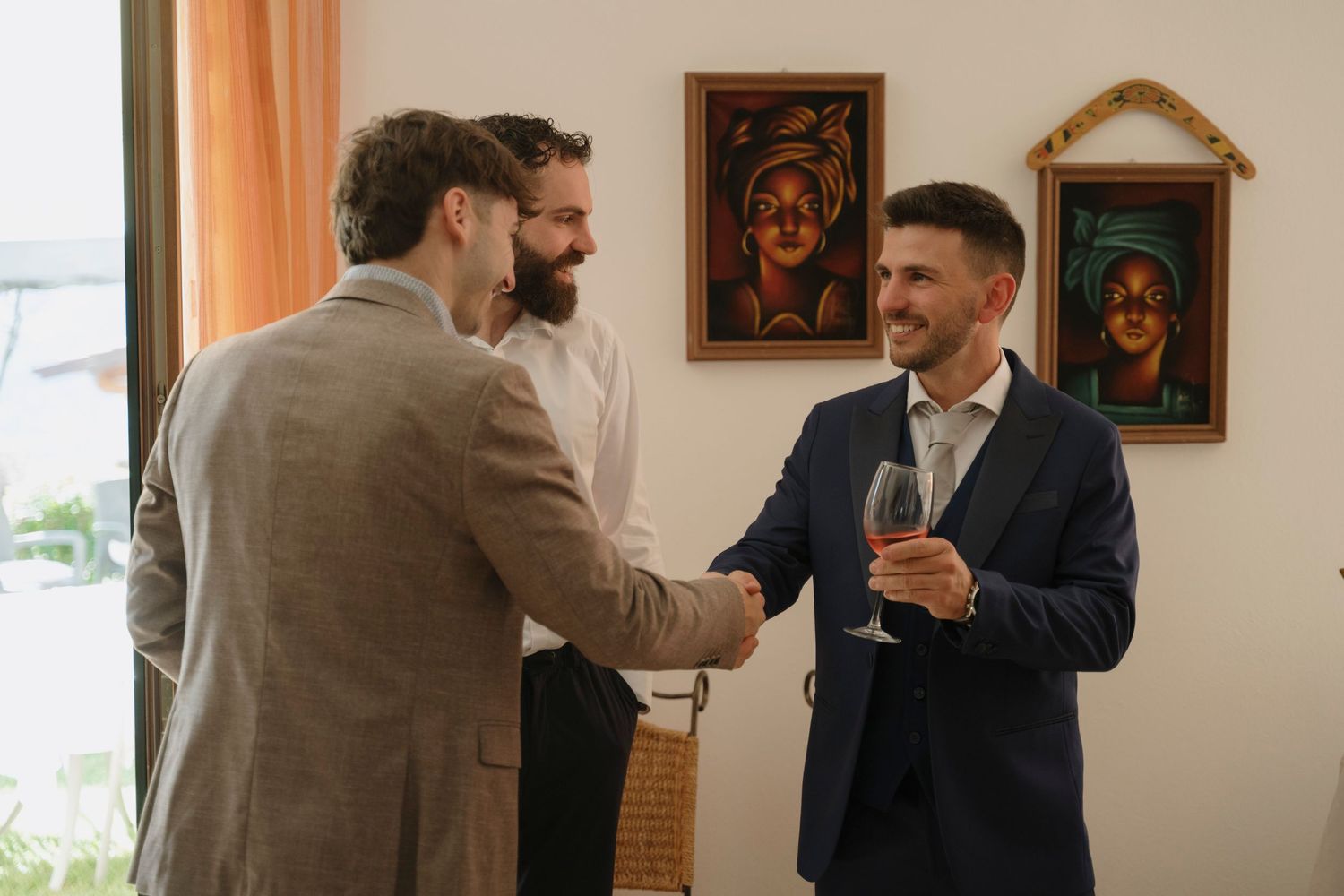 Groom in navy suit shaking hands and greeting guests while holding a glass of wine at wedding reception.
