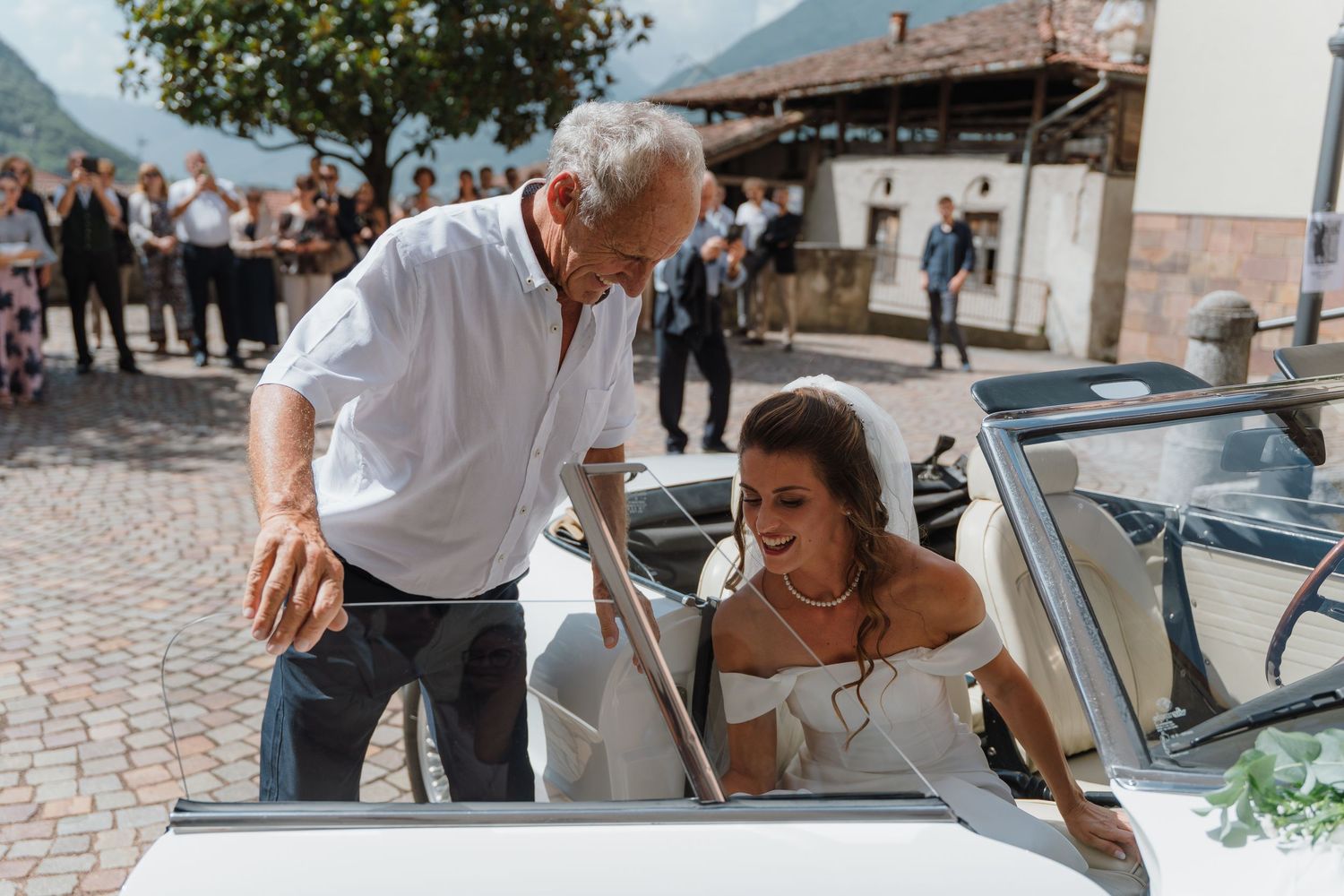 An elderly man helps a bride in a white dress get into a classic white convertible car at an outdoor wedding venue.