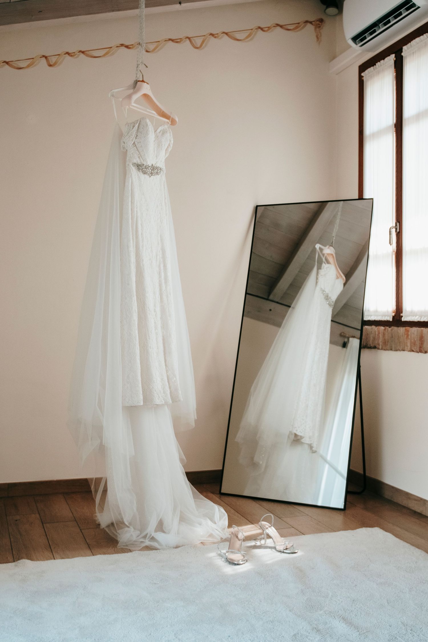 White wedding dress hanging in a bright room with a floor mirror reflection and bridal heels on a white rug.