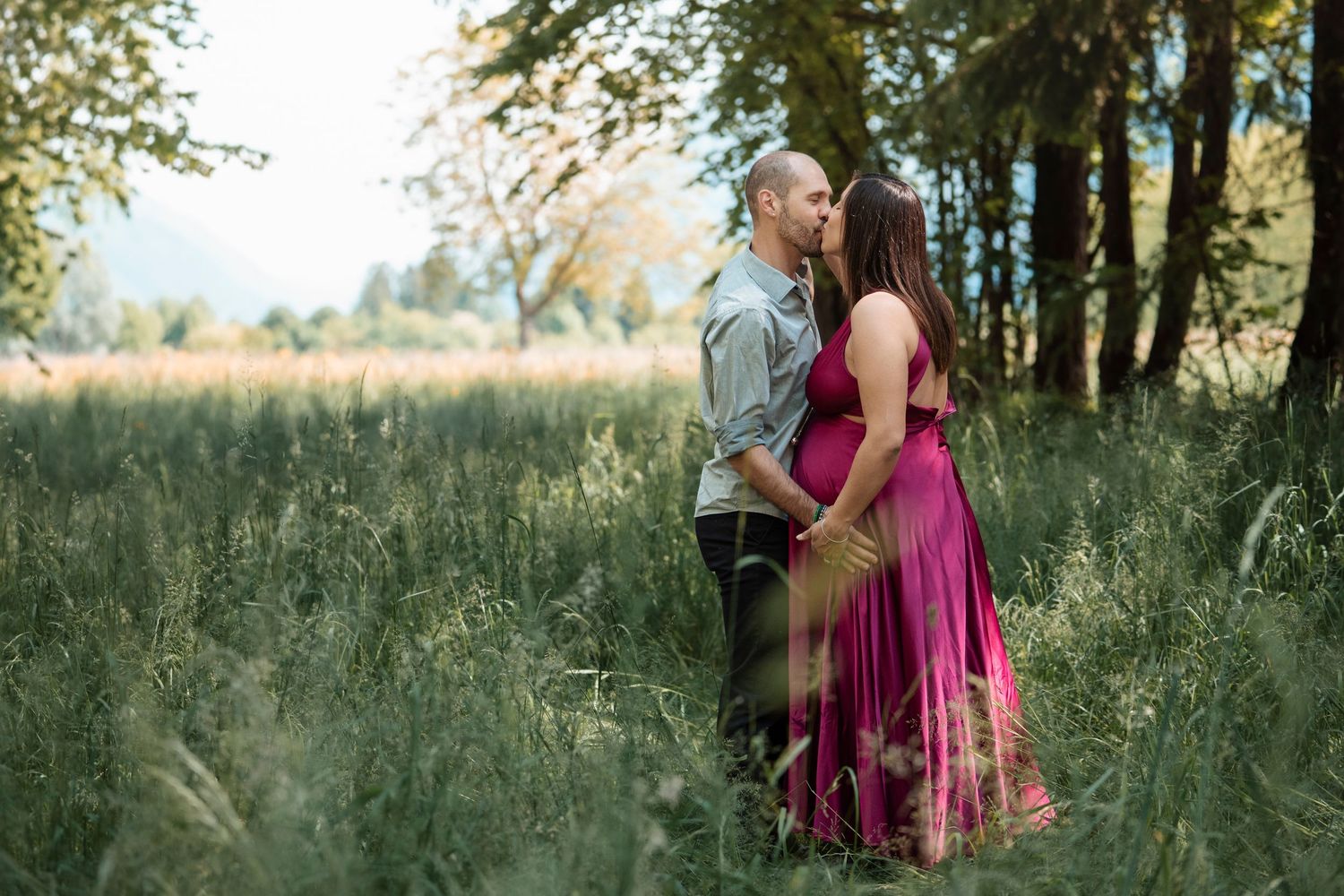 Couple sharing a romantic kiss in a sunlit meadow, woman wearing a flowing pink maternity dress.