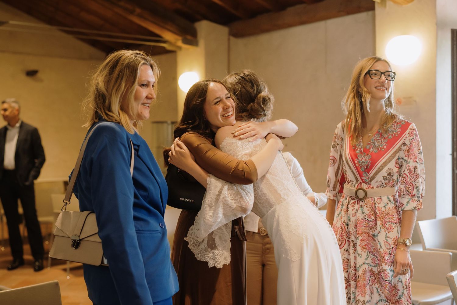 Una sposa in abito di pizzo bianco abbraccia un invitato, mentre alcune donne sorridenti osservano la scena dopo la cerimonia.