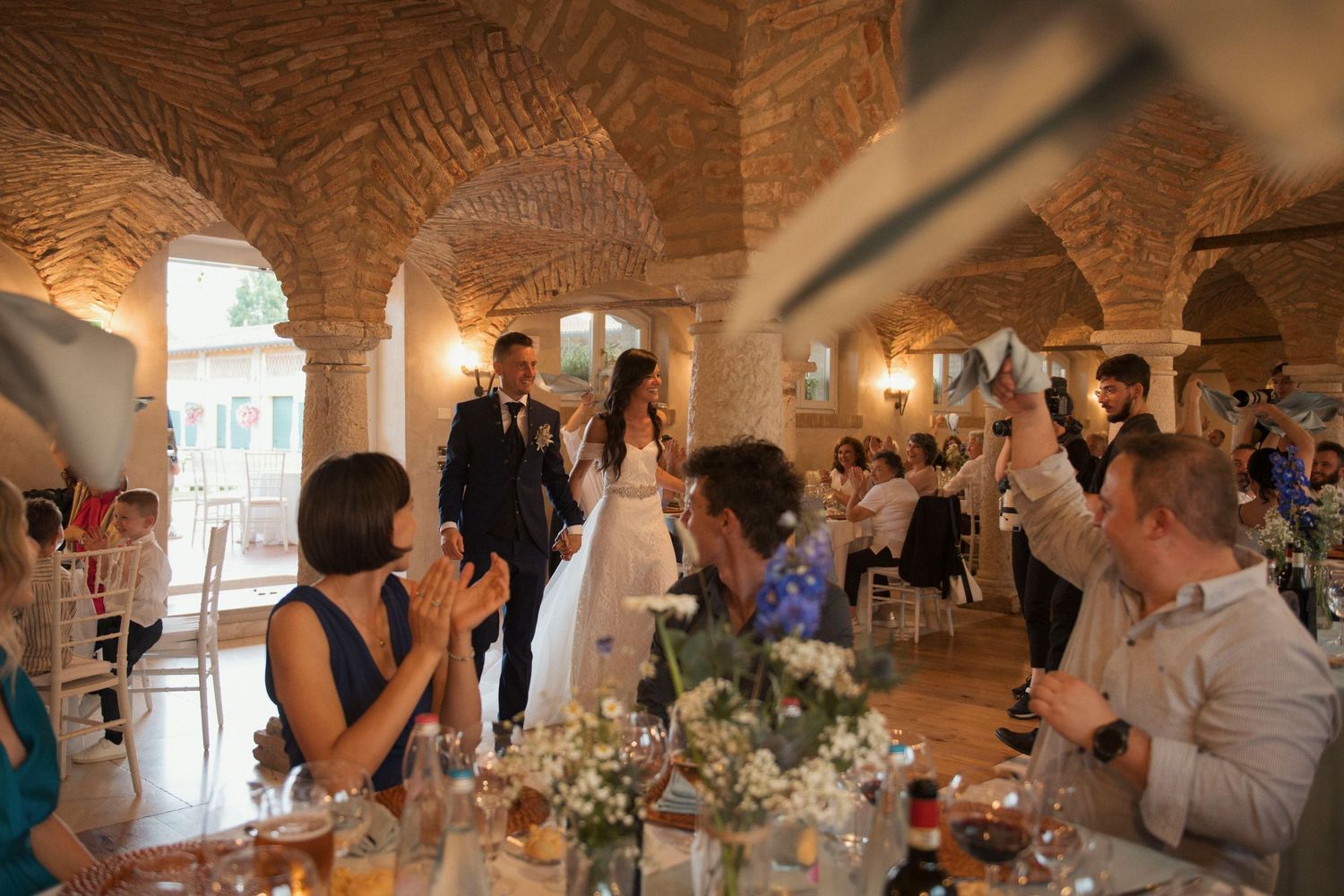 Wedding couple enters reception hall with brick arches as guests applaud and celebrate.