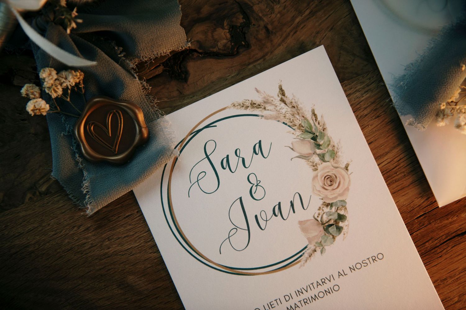 Elegant wedding invitation for Sara and Ioan with floral wreath design, wax seal, and blue ribbon on rustic wood.