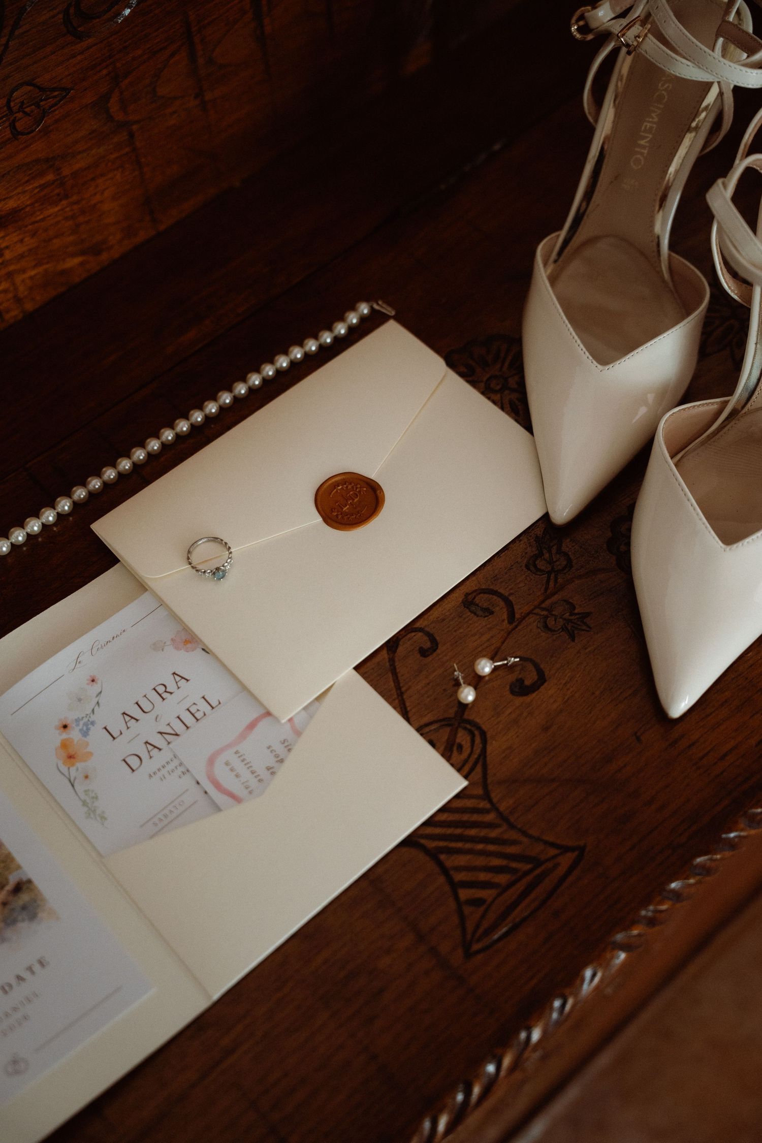 Bridal flat lay featuring white heels, pearl necklace, wedding invitation, wax seal envelope, engagement ring, and earrings on wood.
