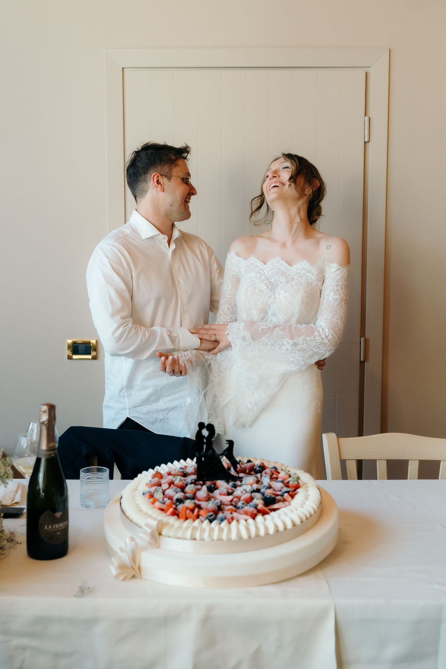 Laughing bride and groom in white attire prepare to cut decorated wedding cake together.