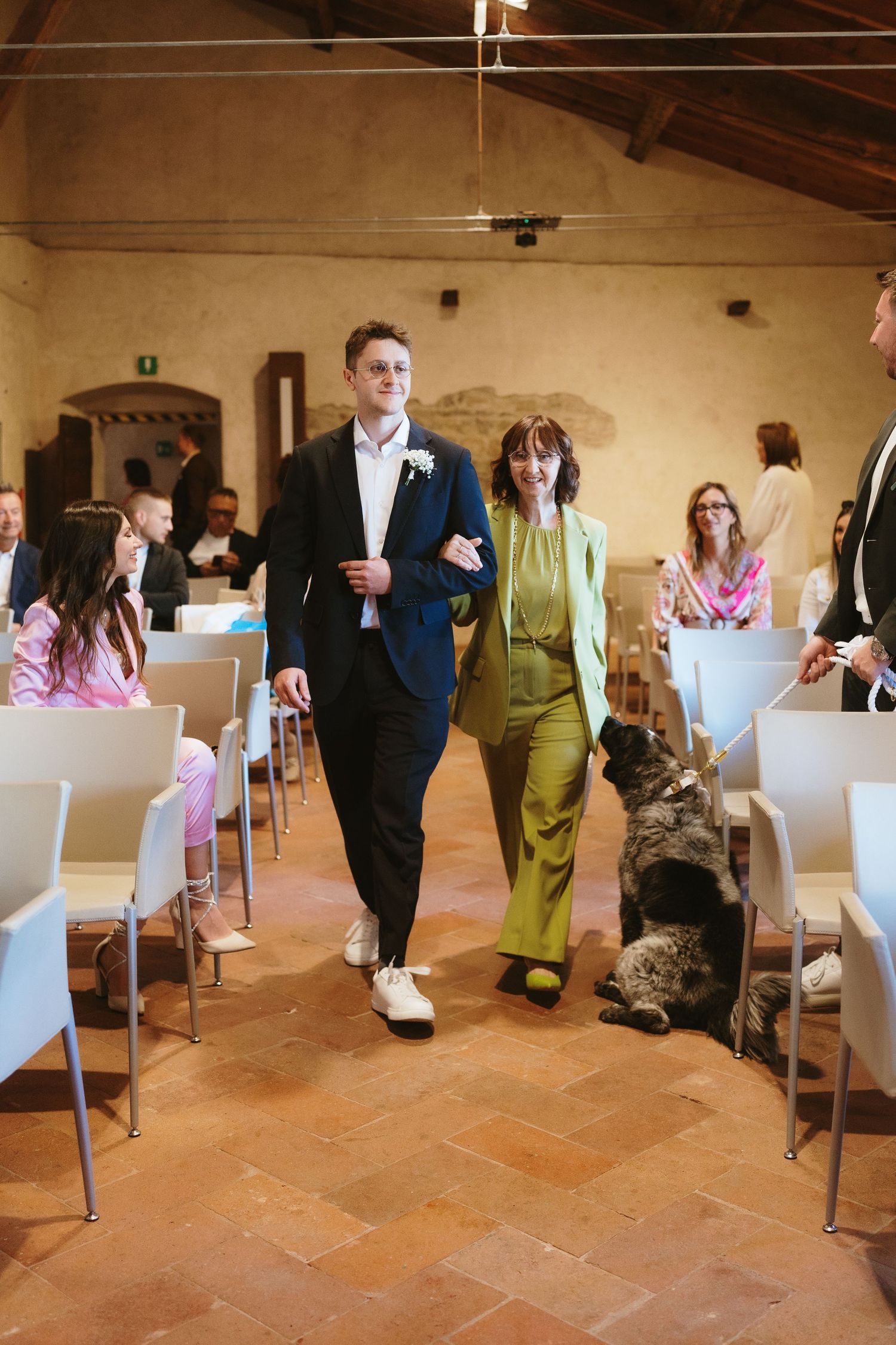 Groom in navy suit walks down aisle with woman in green pantsuit at rustic Italian wedding venue.