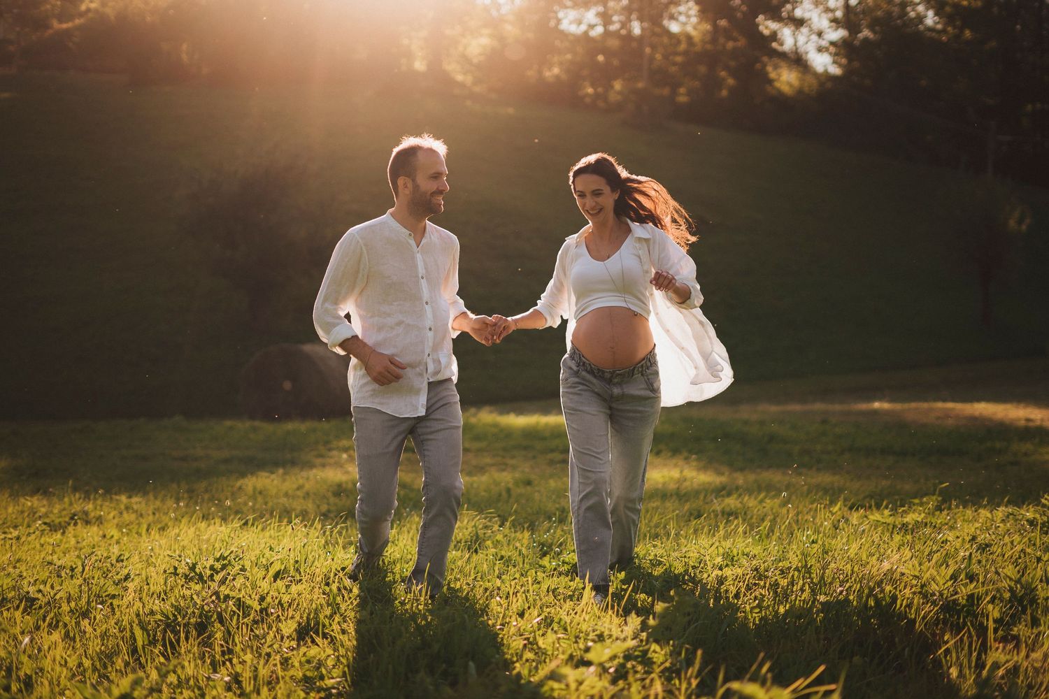 Joyful expecting couple running hand in hand through a golden sunlit green field at sunset.