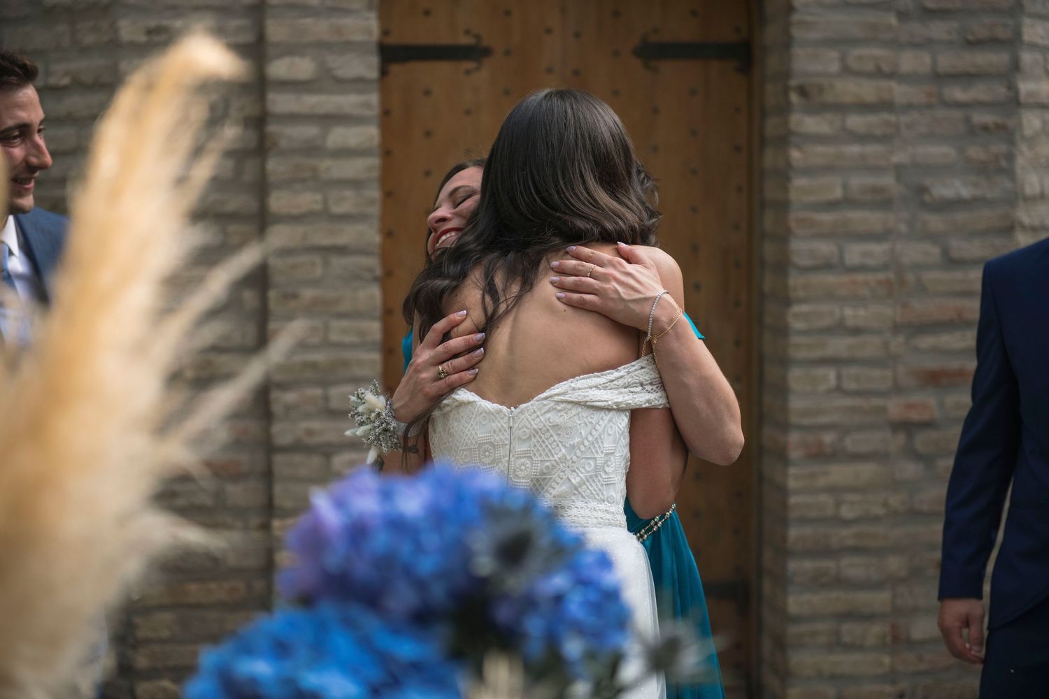 Bride in off-shoulder wedding gown shares heartfelt hug with guest near brick wall at reception.