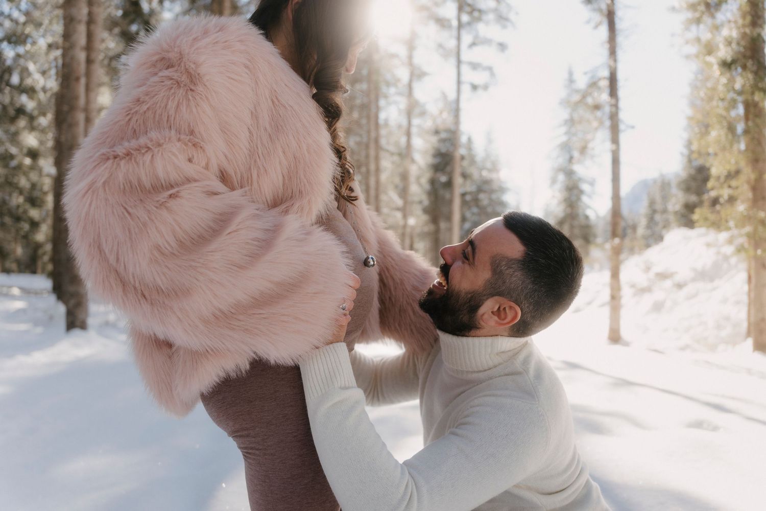 Man kneeling in snow kissing pregnant woman's baby bump in a sunny winter pine forest.