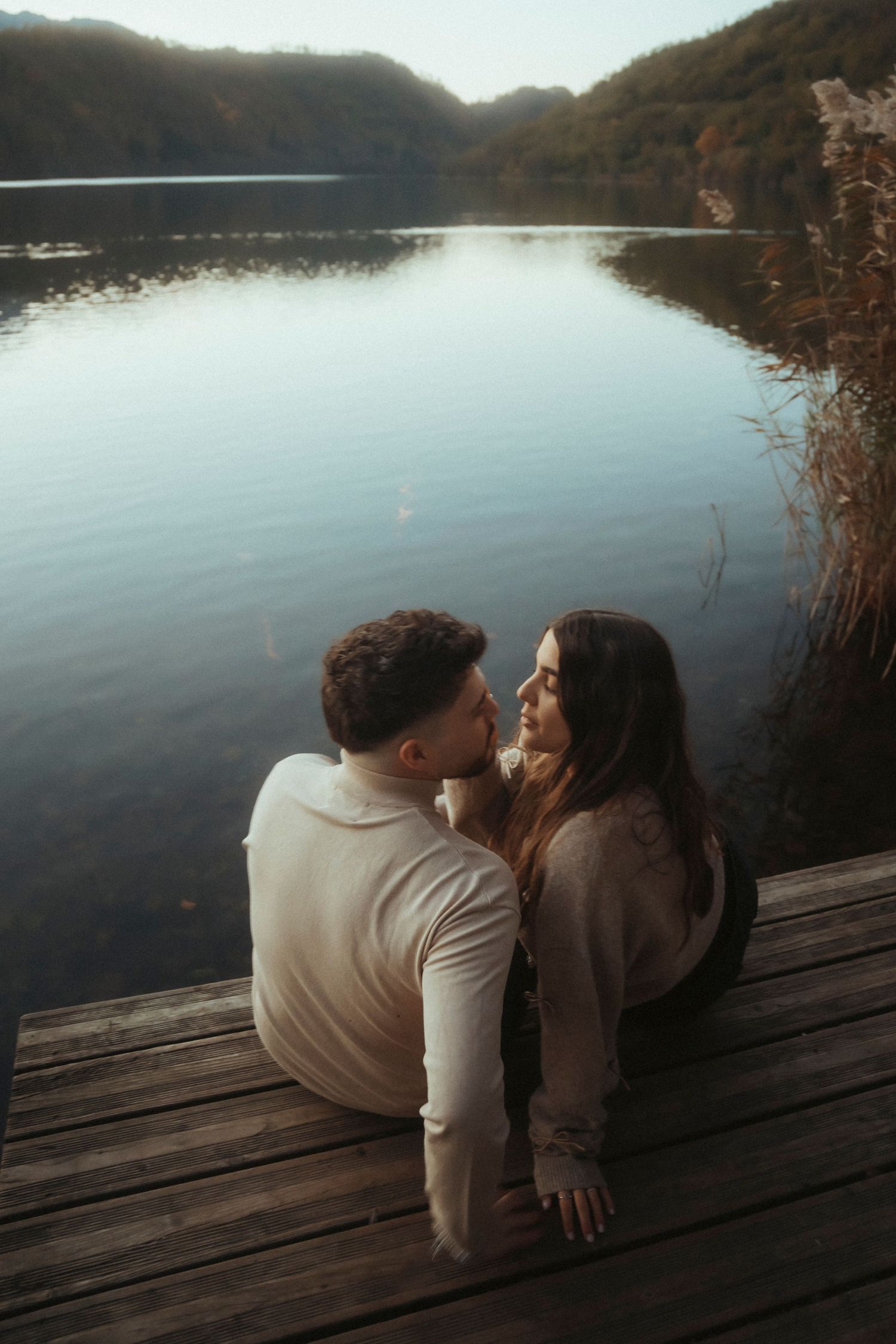 Romantic couple sitting close on a wooden dock overlooking a calm mountain lake.