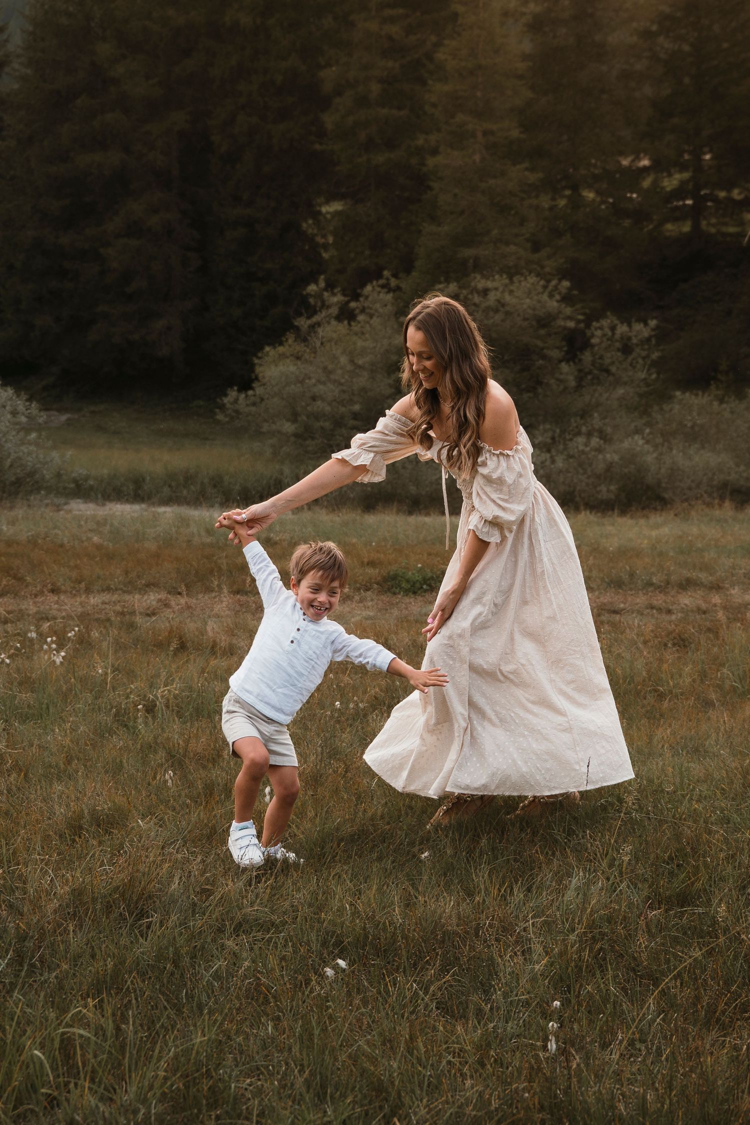Joyful mother in cream dress dancing with young son in a lush green meadow.