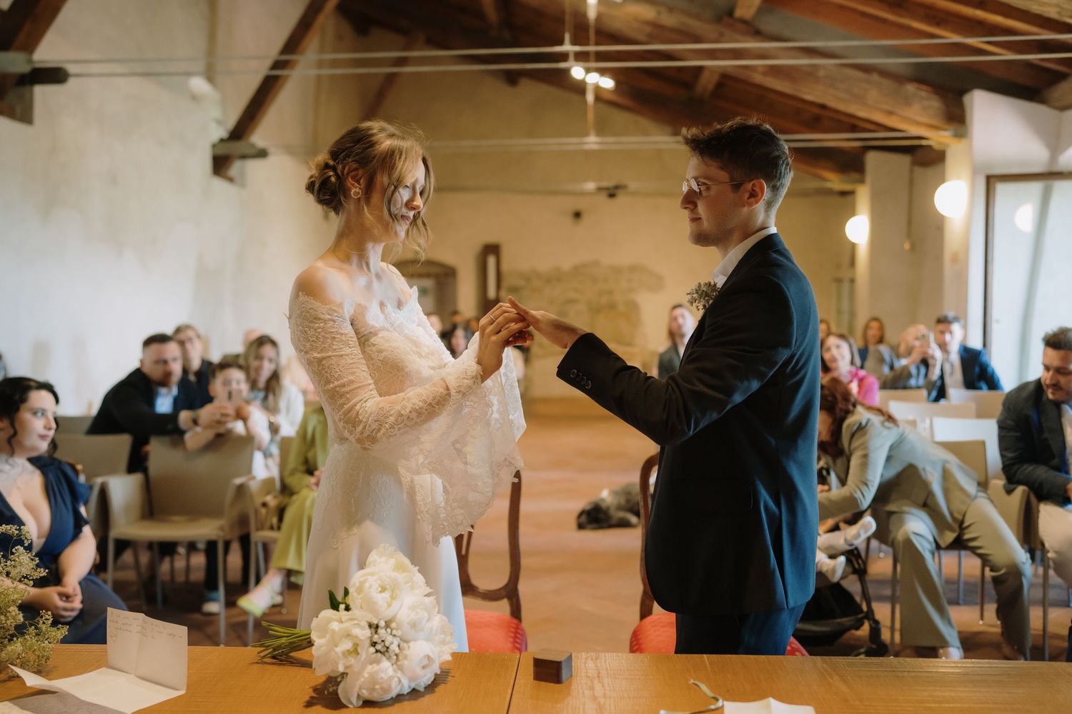 Bride and groom exchange rings during an intimate wedding ceremony in a rustic venue with seated guests.