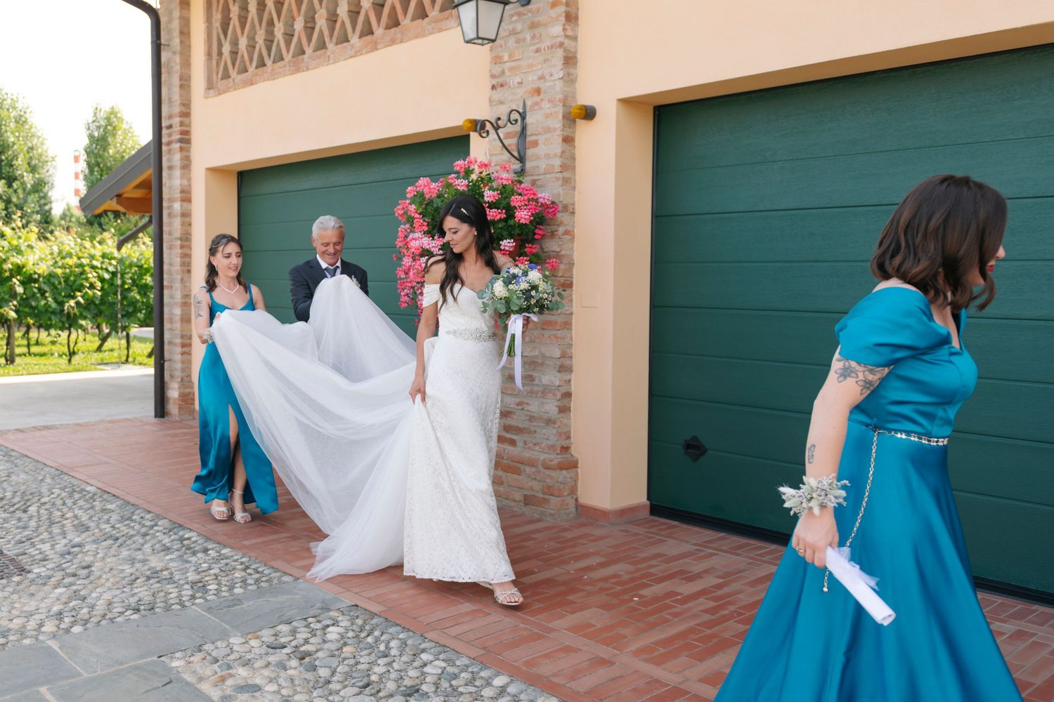 Bride in white gown with bridesmaids in teal dresses walking outside a rustic venue adorned with pink flowers.