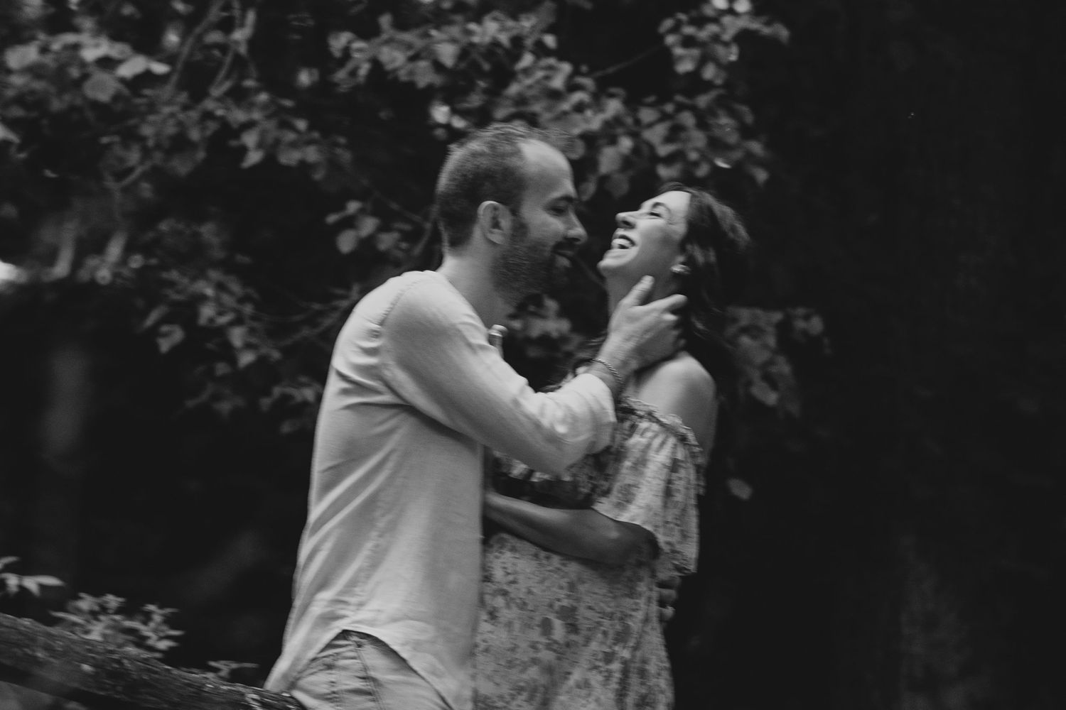 Black and white photo of a laughing couple embracing in a lush forest setting.