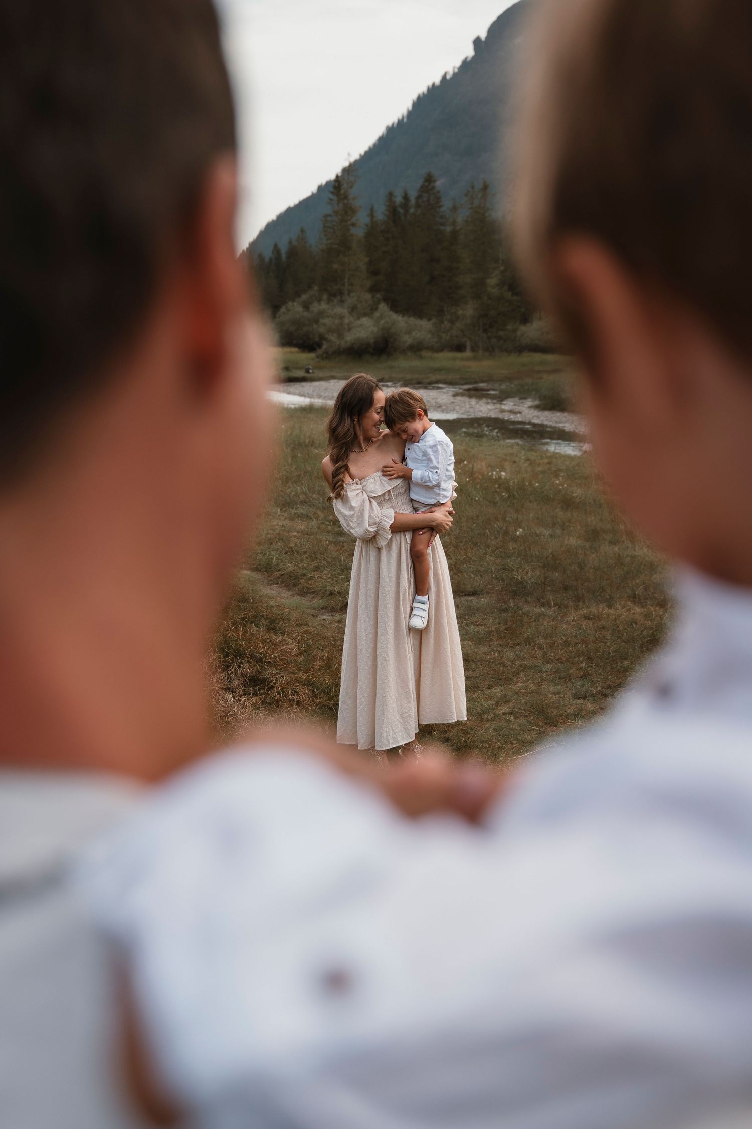 A mother in a white dress holds her young child in a scenic mountain meadow while two people watch in the foreground.