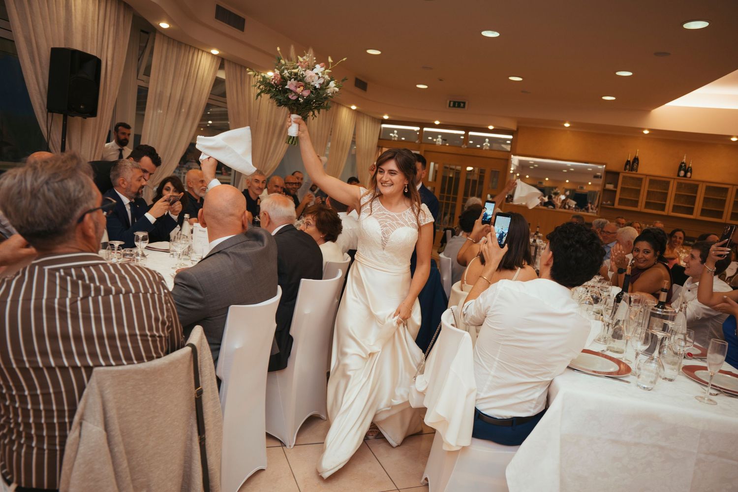 Bride in white dress raising her bouquet while making a grand entrance at the wedding reception dinner.