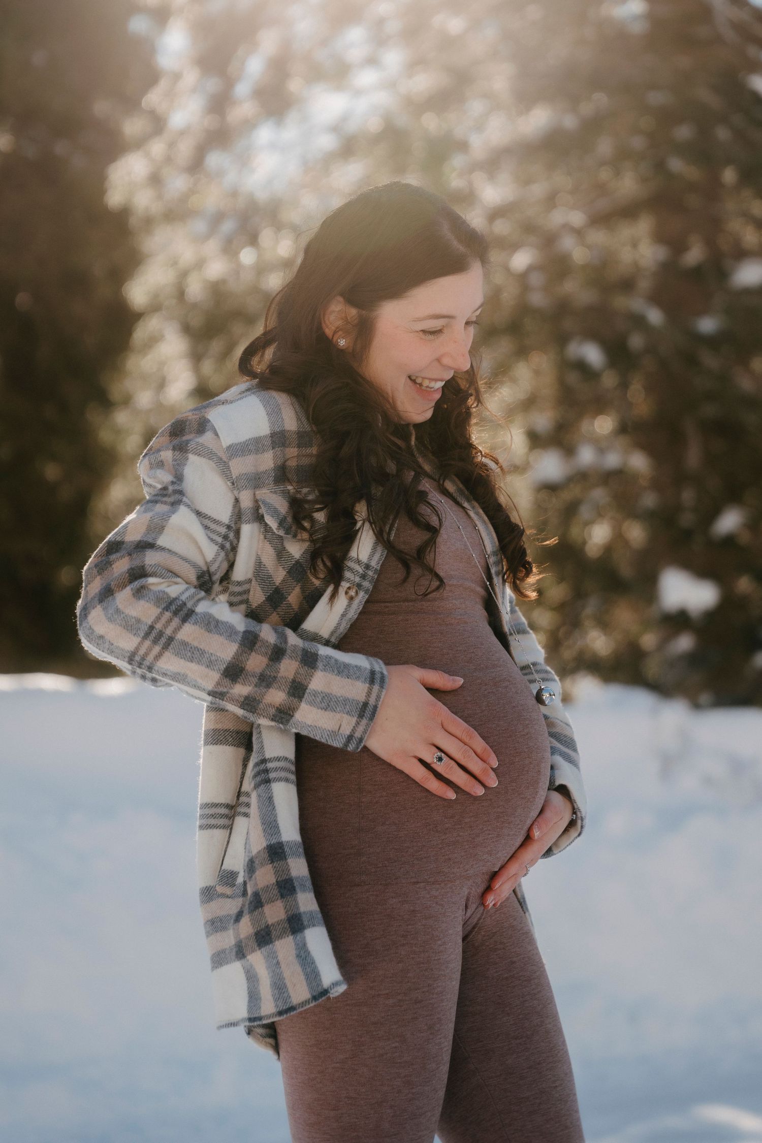 Smiling pregnant woman in plaid shirt cradling baby bump outdoors in a sunny snowy winter setting.