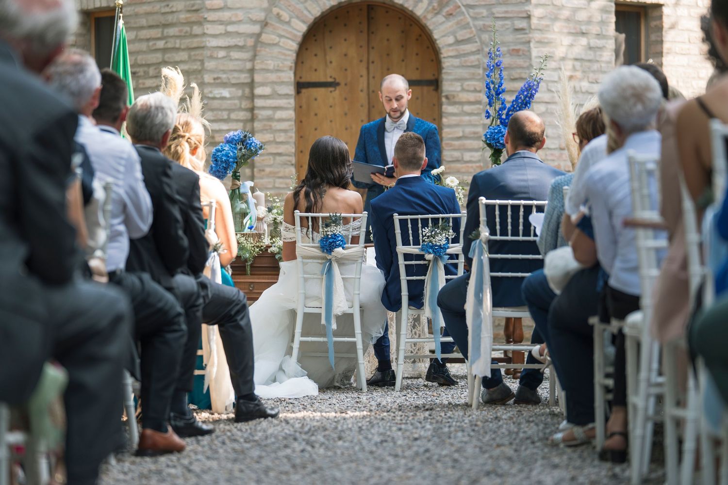 Outdoor wedding ceremony with guests seated as officiant reads vows at rustic stone venue.