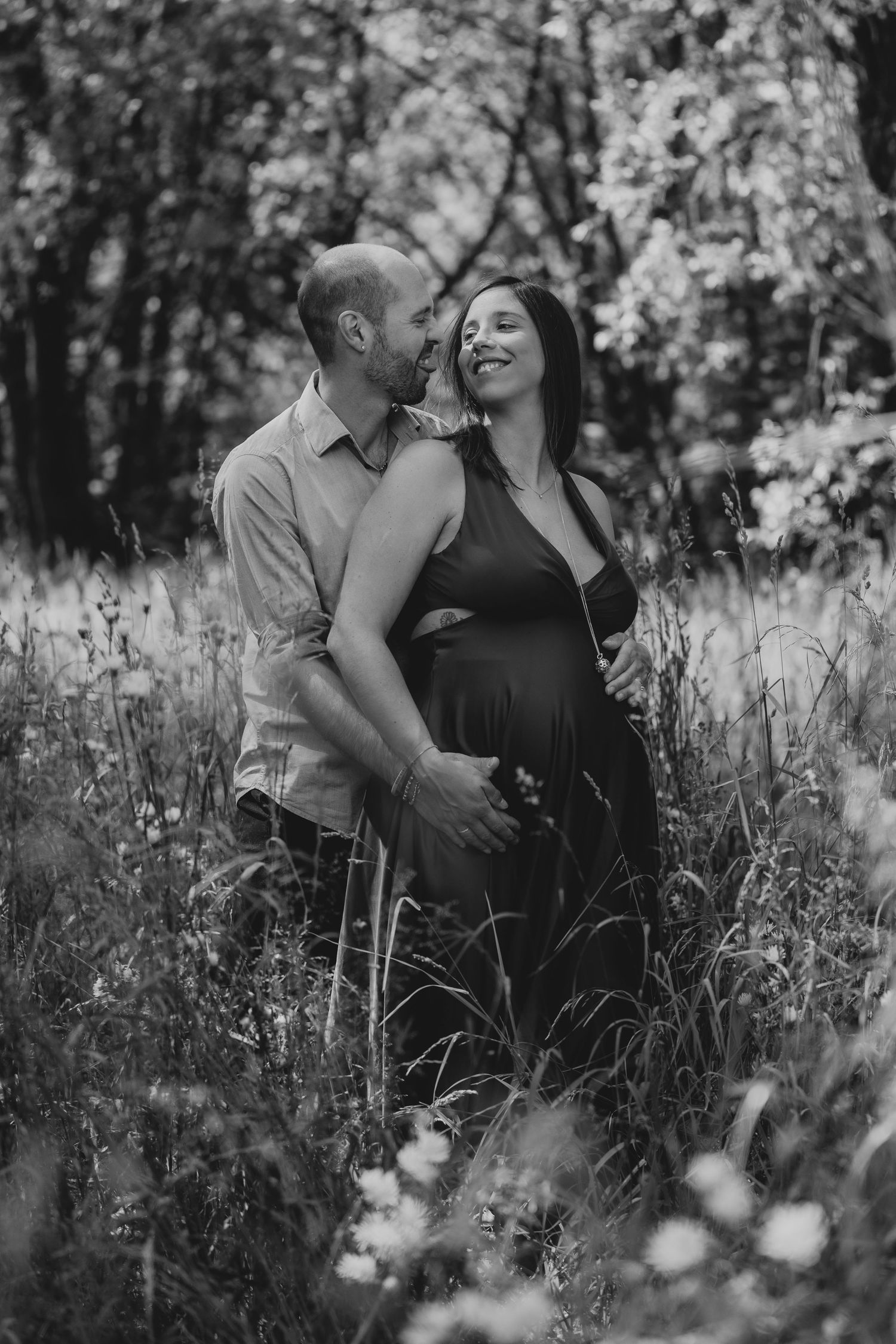 Black and white photo of a couple embracing in a wildflower meadow, woman visibly pregnant.