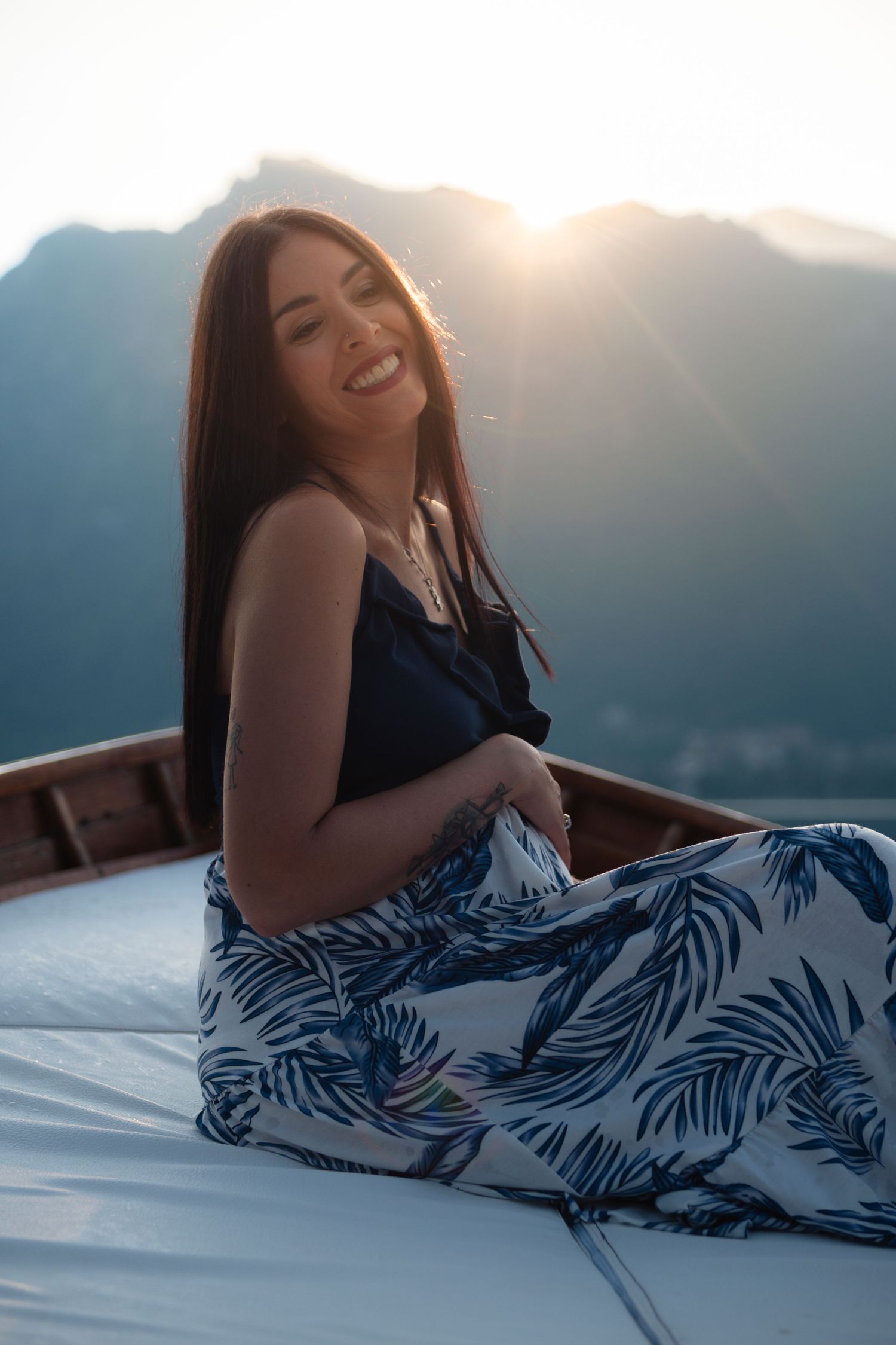 Smiling young woman in floral skirt sitting on a boat with mountain sunset backdrop.