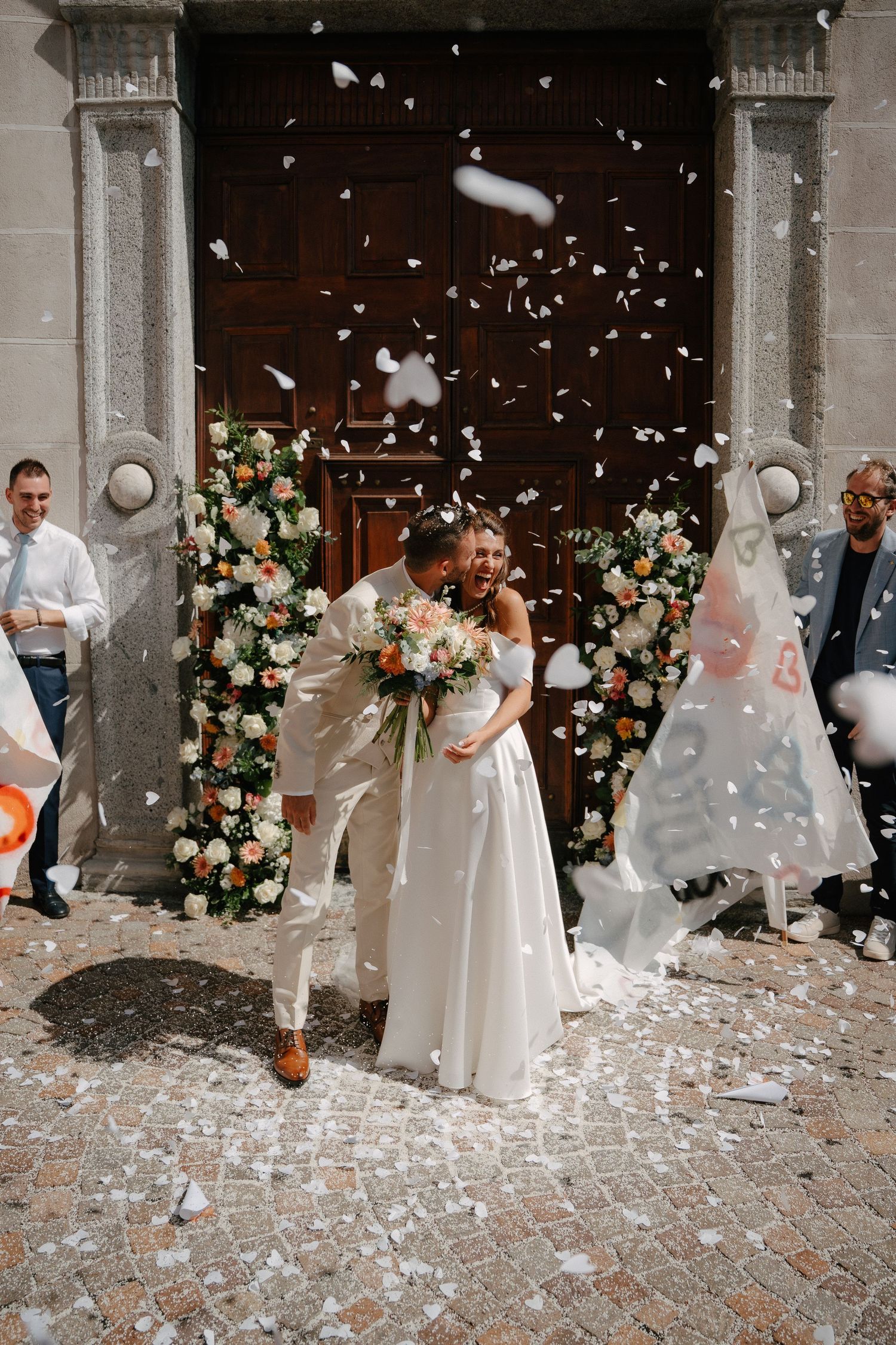 Bride and groom kissing outside church entrance with white confetti falling, floral arch backdrop.