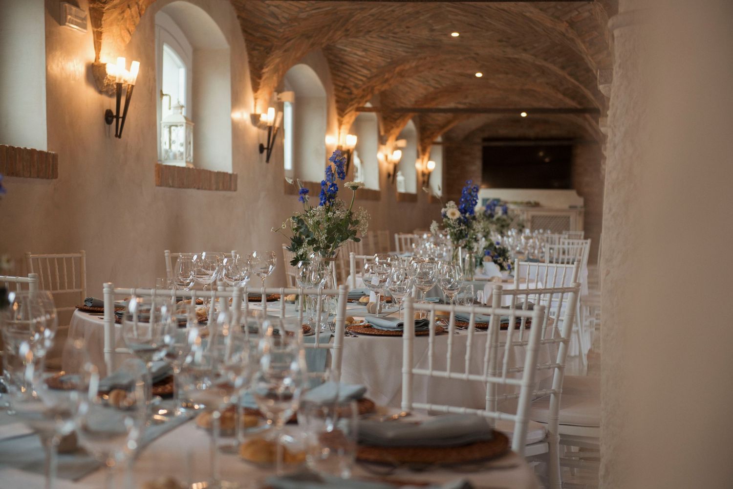 Elegant wedding reception in a vaulted stone venue with white Chiavari chairs, floral centerpieces, and candlelit tables.