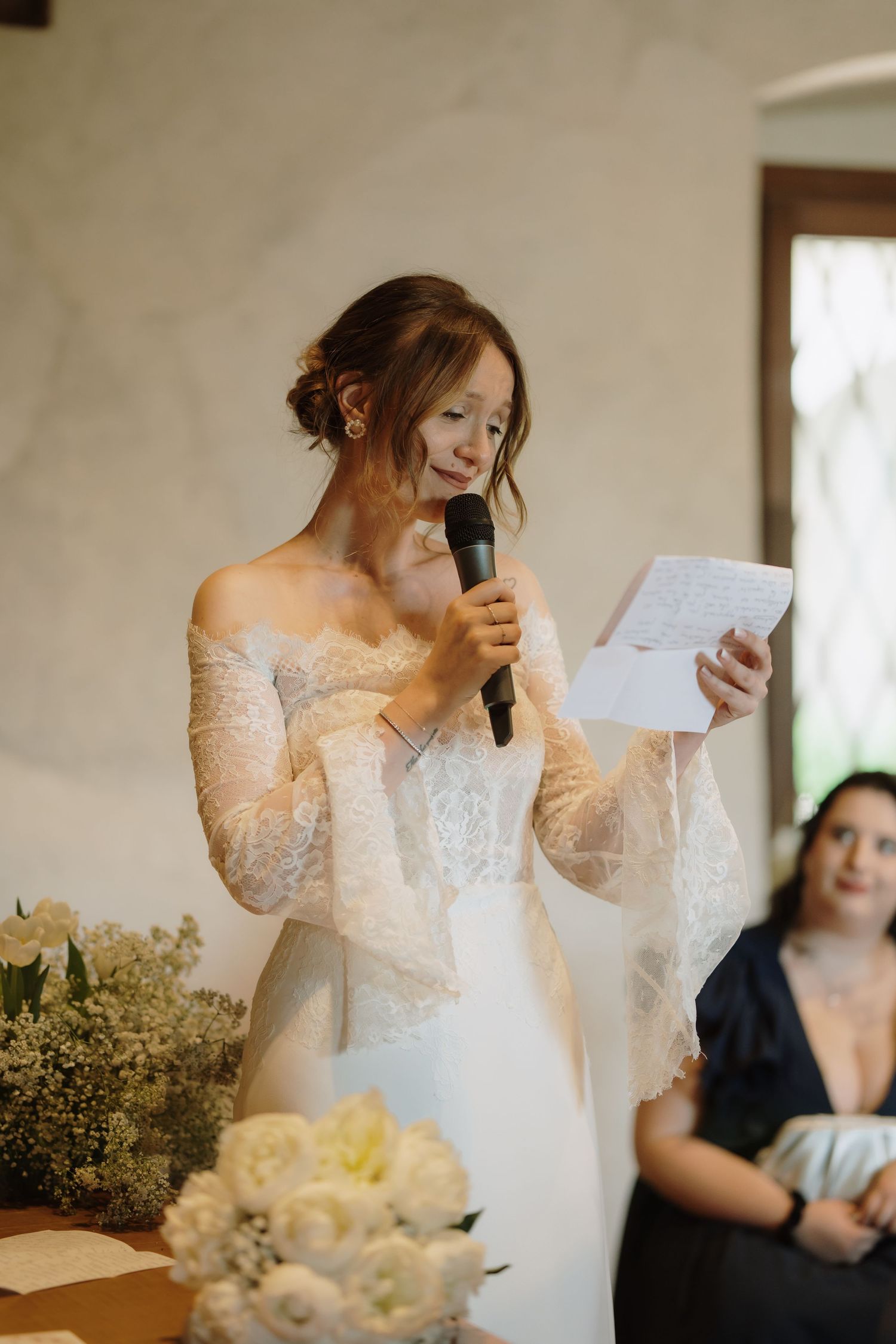 Emotional bride in lace off-shoulder gown reads vows into microphone holding paper at wedding ceremony.