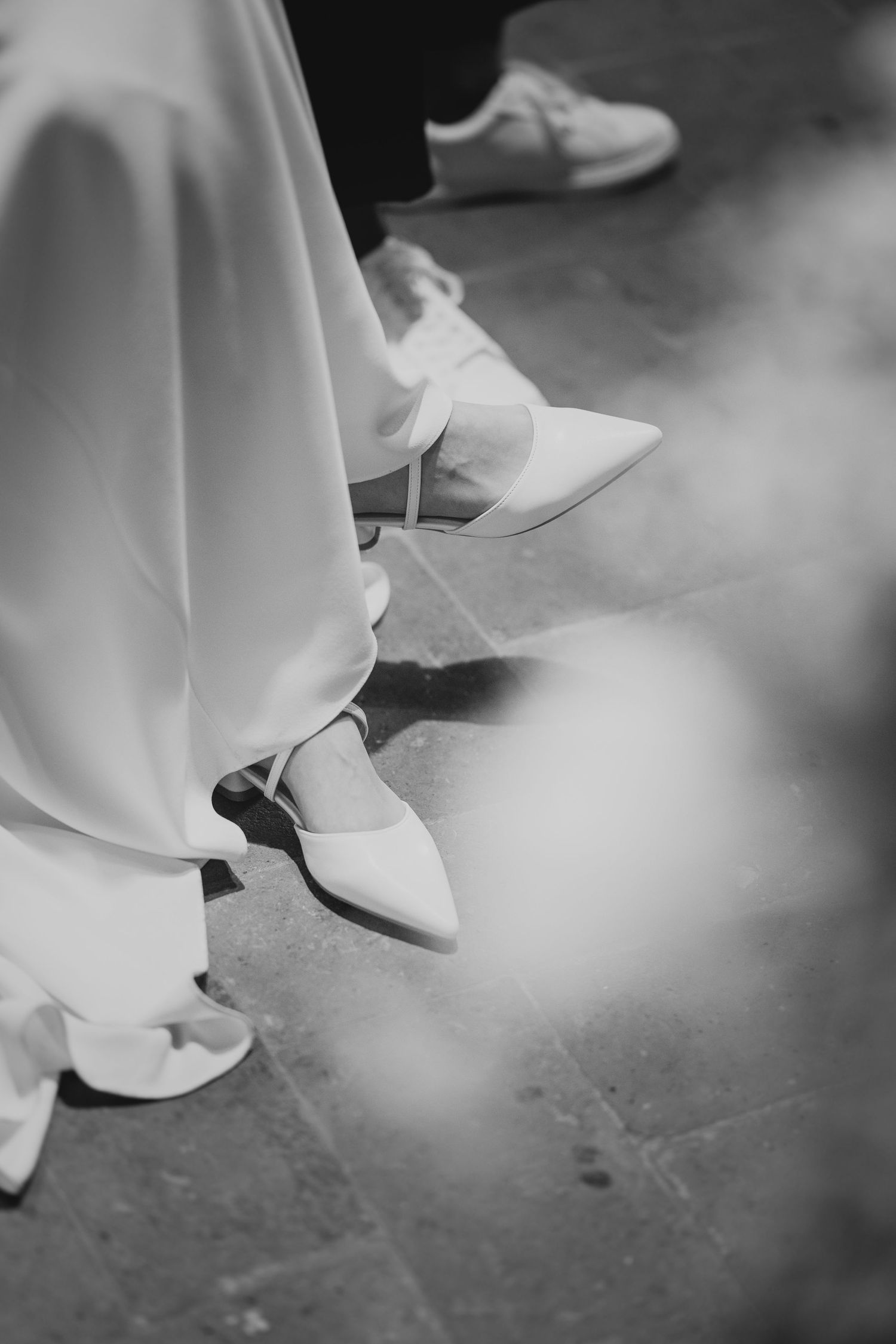 Black and white close-up of bride's white pointed heels beneath a flowing wedding dress hem.
