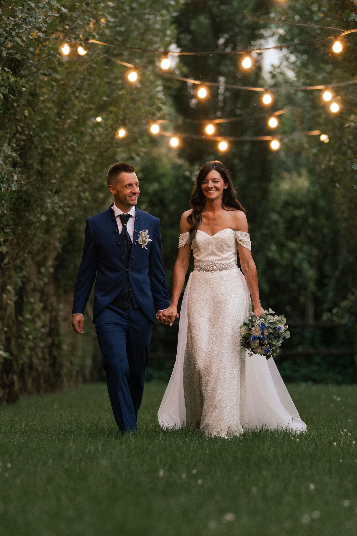 Bride and groom walk hand-in-hand on green grass under glowing string lights surrounded by lush trees at dusk.