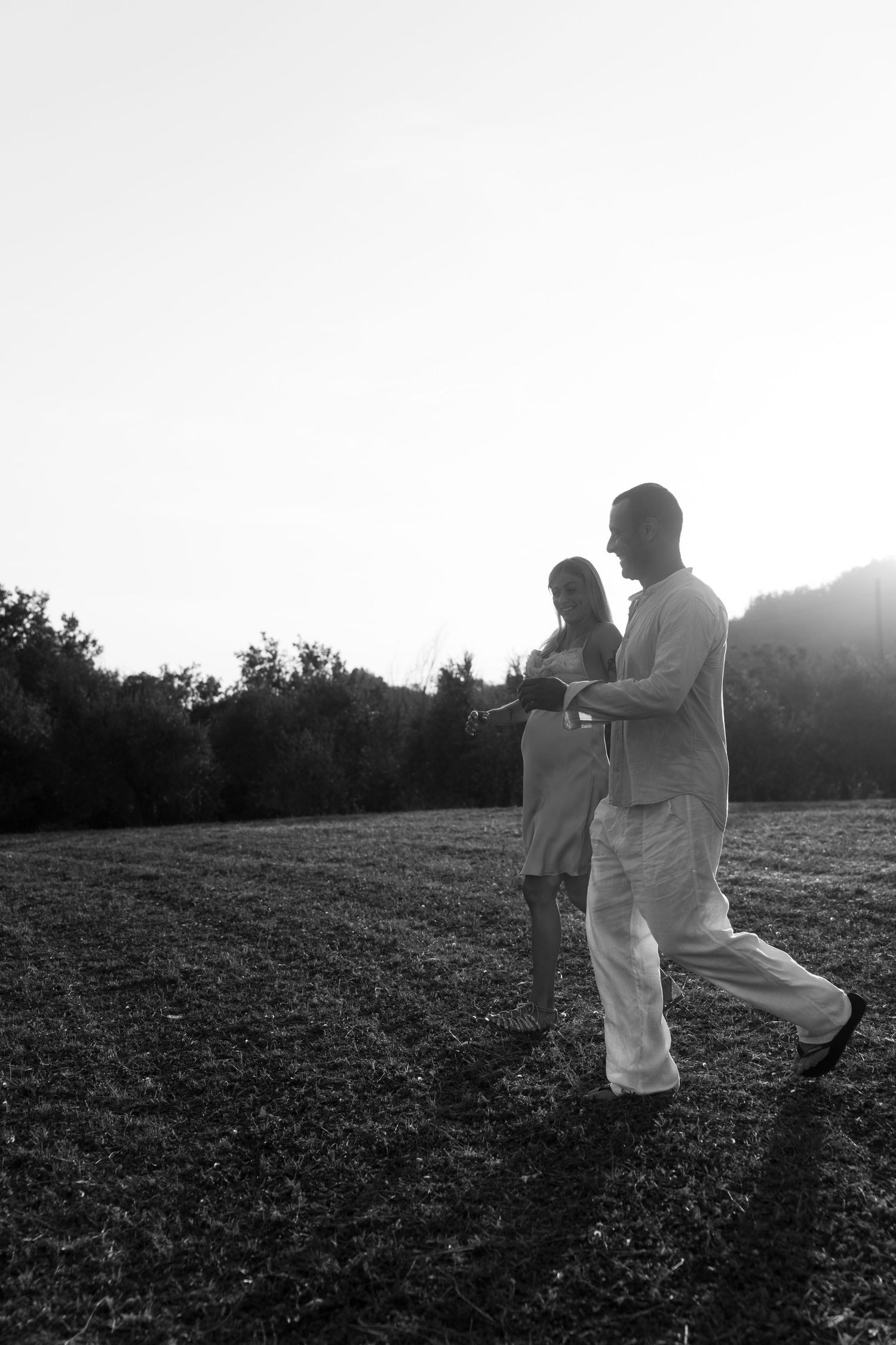 Black and white photo of a couple walking hand in hand through a sunlit field with trees in the background.