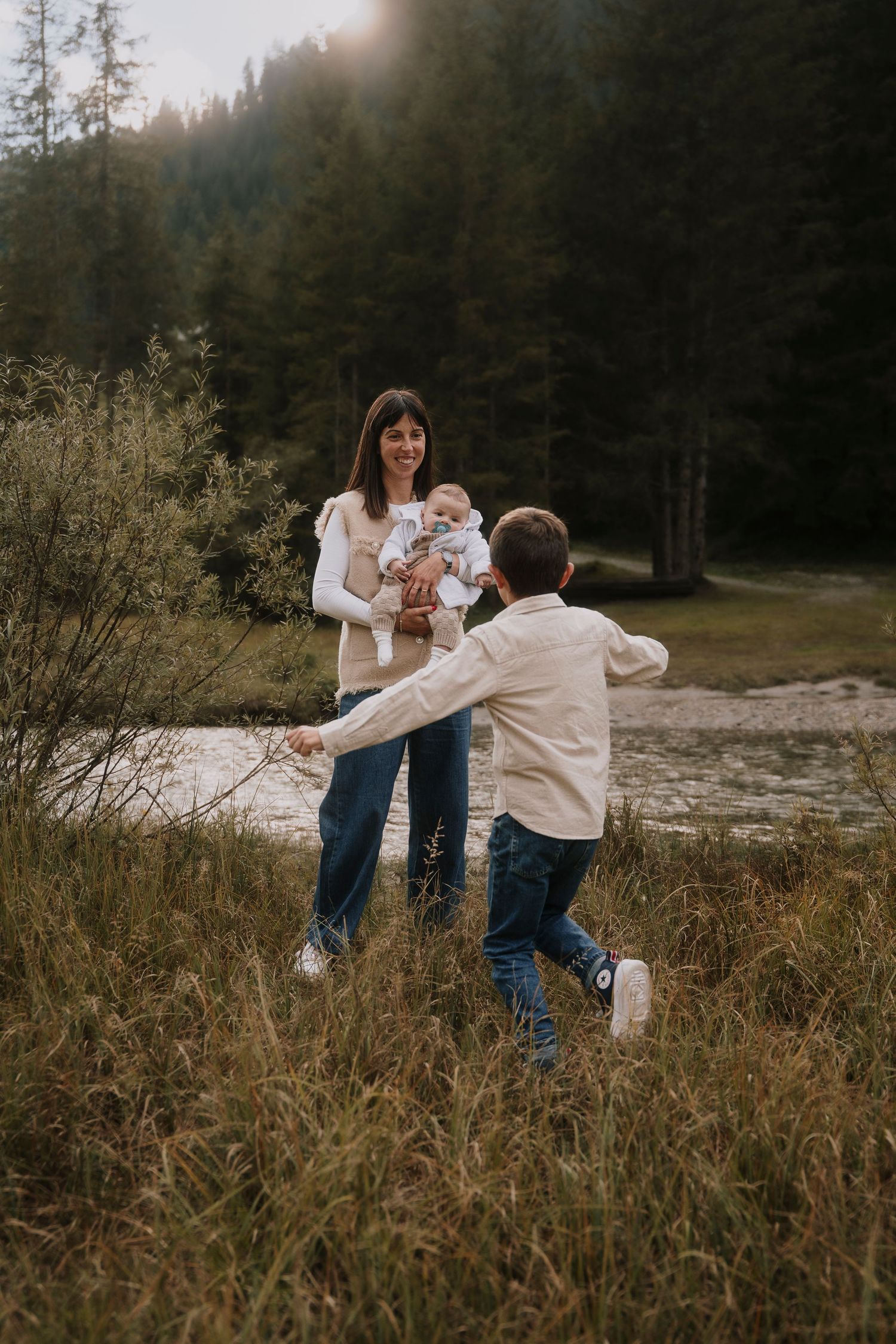 Mother carrying baby in carrier smiles as young son runs toward her in a scenic forest meadow.