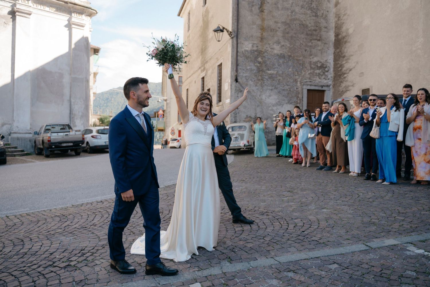 Newlywed couple walking out of church as wedding guests line up to celebrate on cobblestone square.