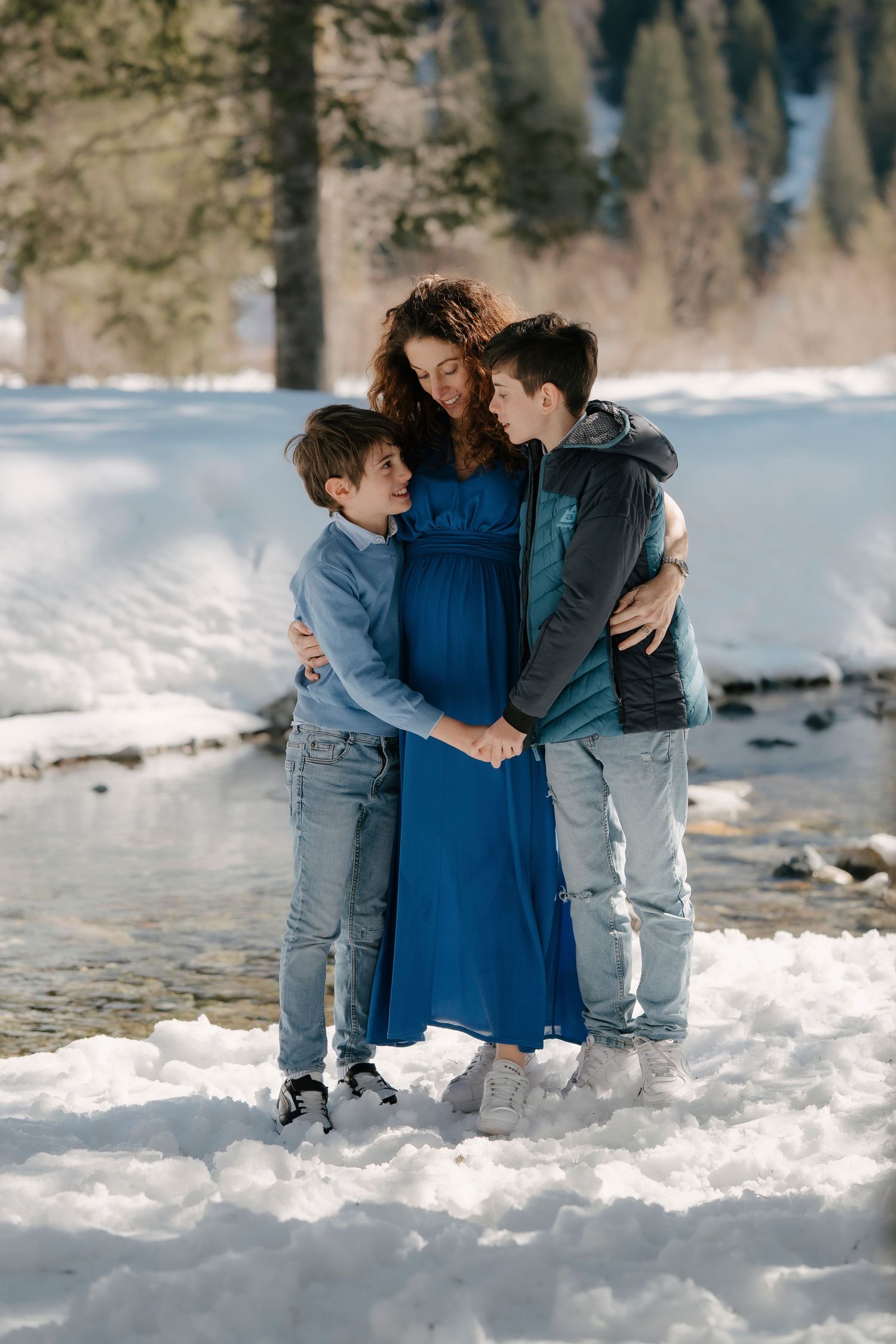 Pregnant woman in blue dress with two boys embracing near snowy river in winter.
