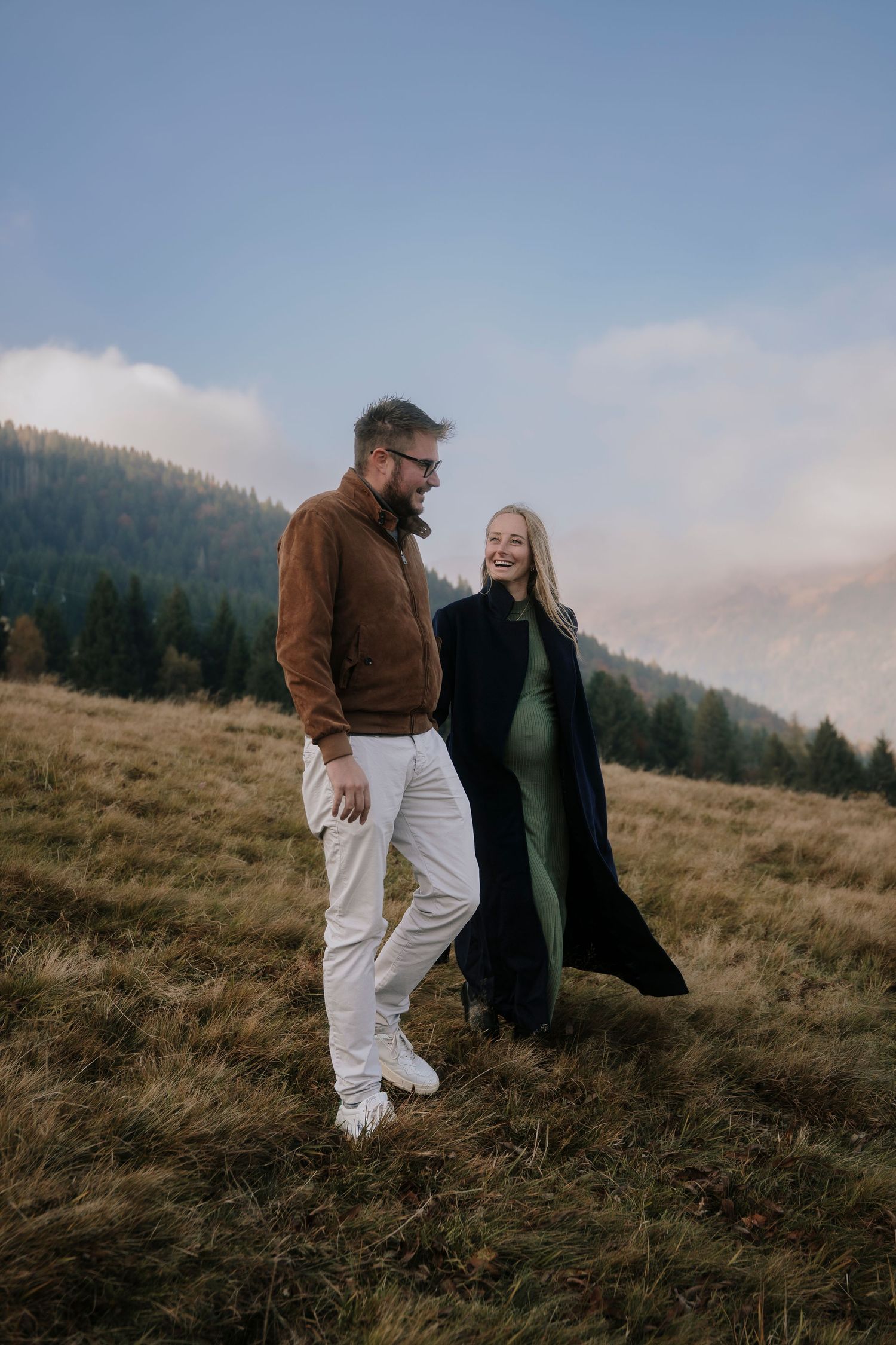 A smiling pregnant couple walks together on an autumn mountain meadow under a cloudy sky.