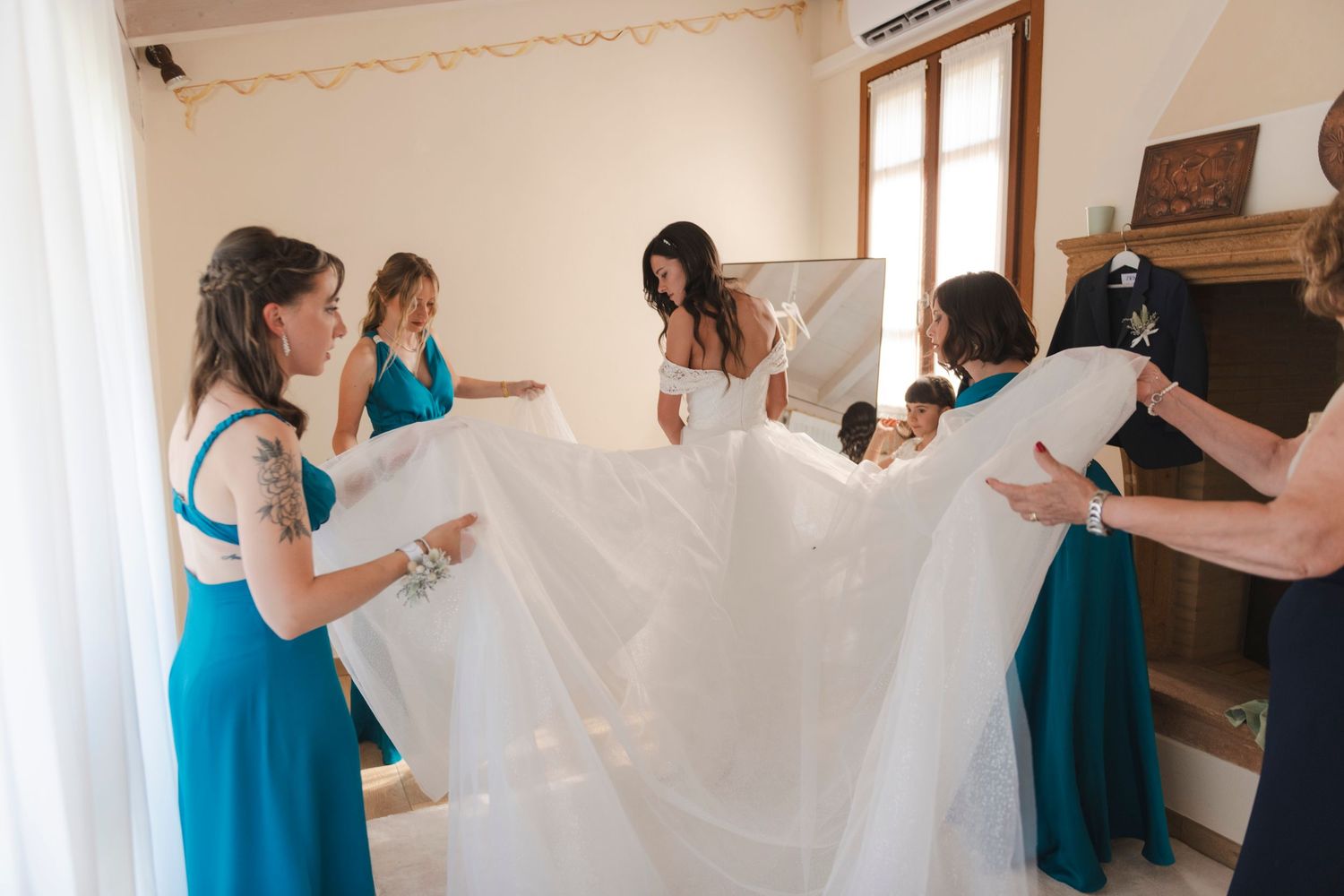 Bride surrounded by bridesmaids in teal dresses helping arrange white wedding gown indoors.