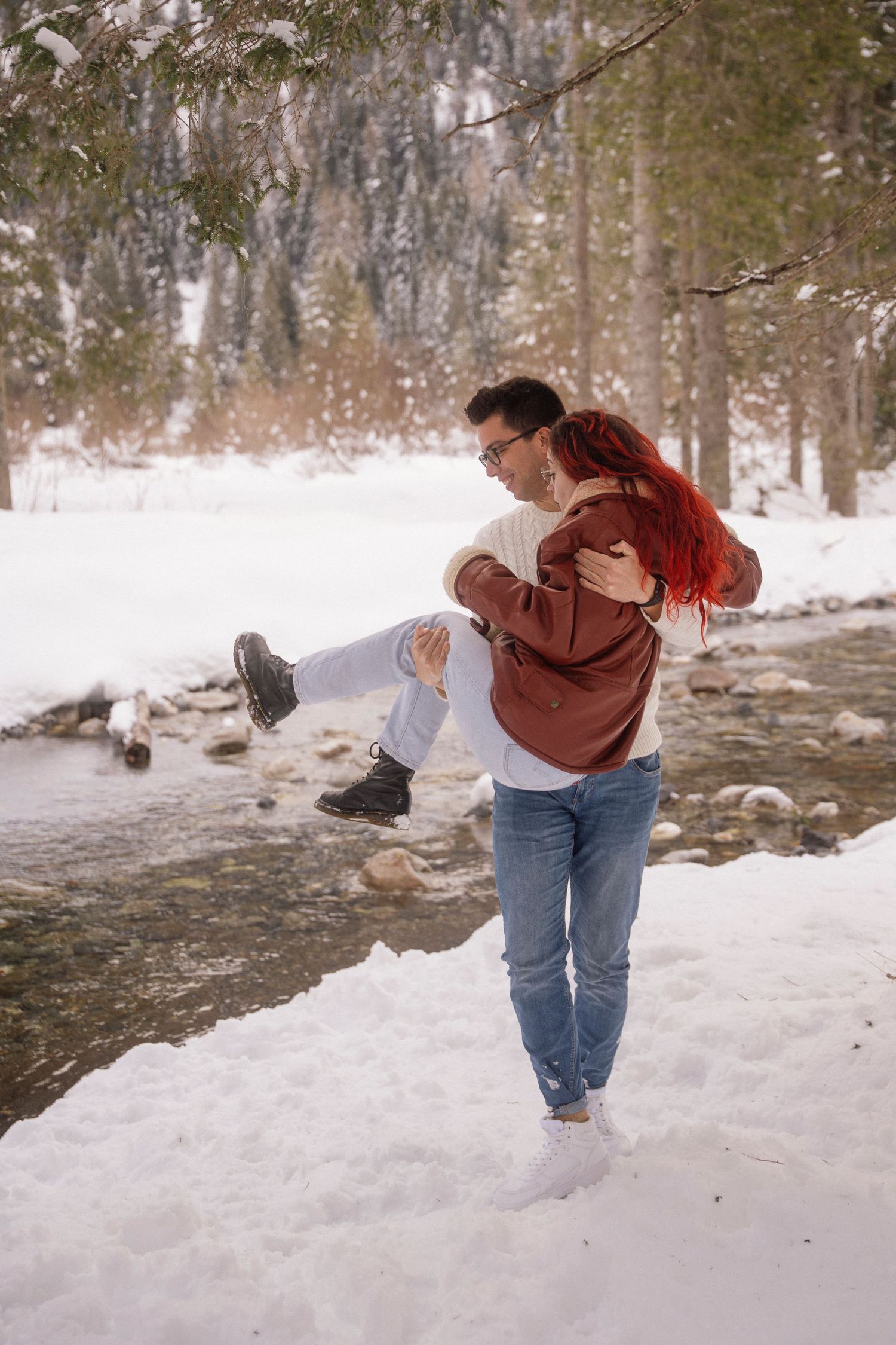 A man in a white sweater and jeans lifts and carries a woman in a red jacket near a snowy riverbank in a winter forest.