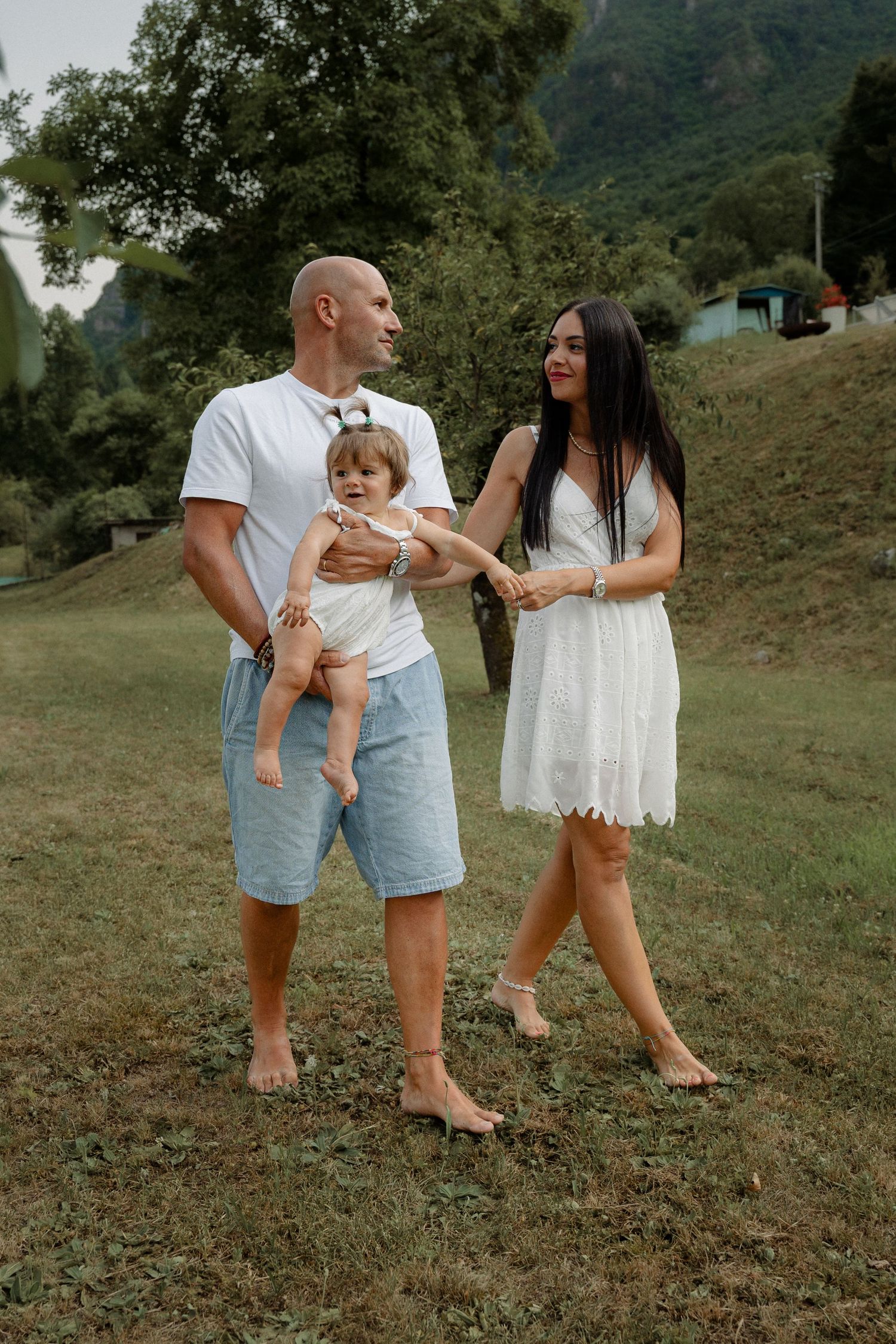 A bald man carries a baby while walking with his smiling dark-haired wife on a grassy hillside path.
