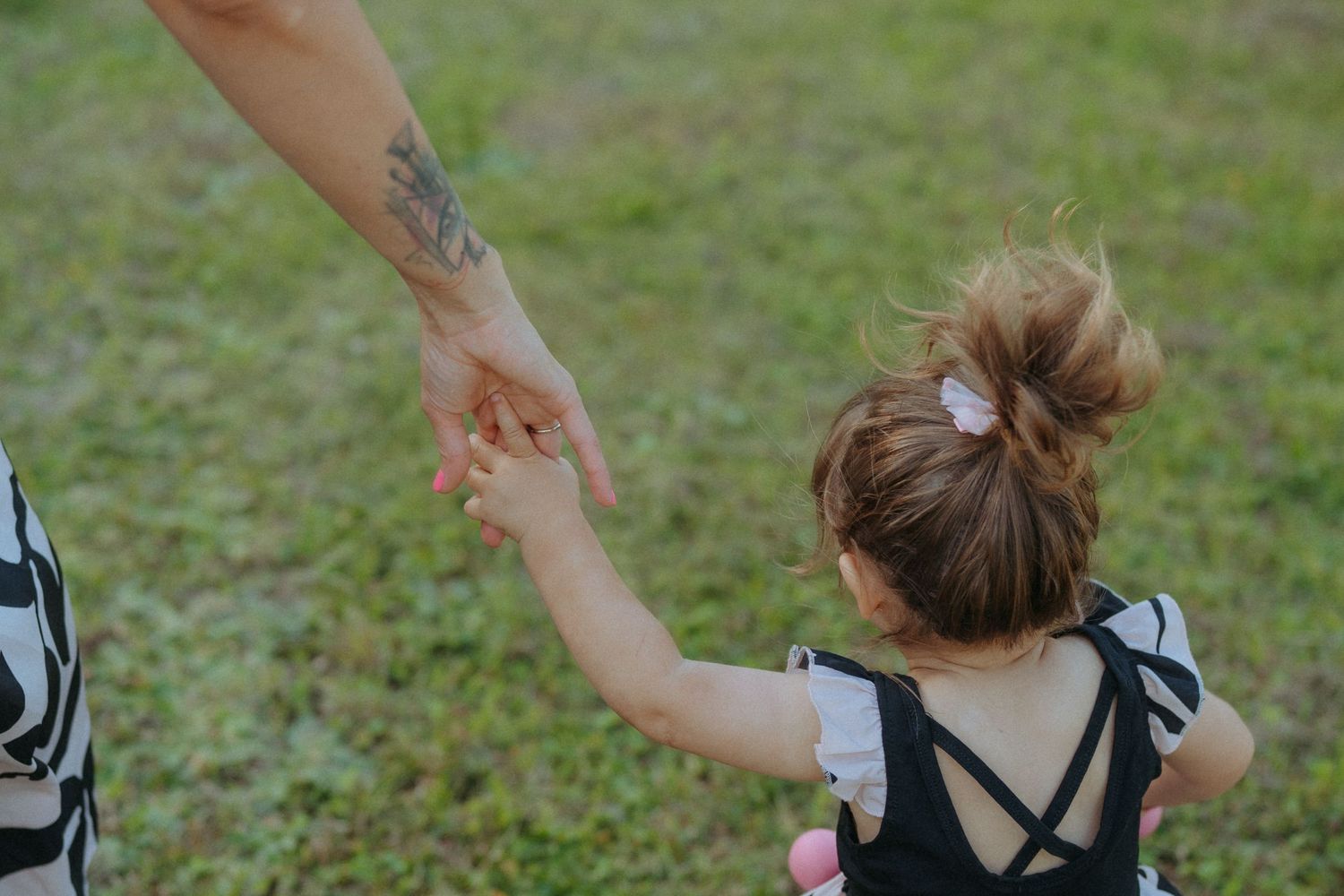 Genitore tatuato tiene per mano una bambina piccola mentre cammina sull'erba verde all'aperto.