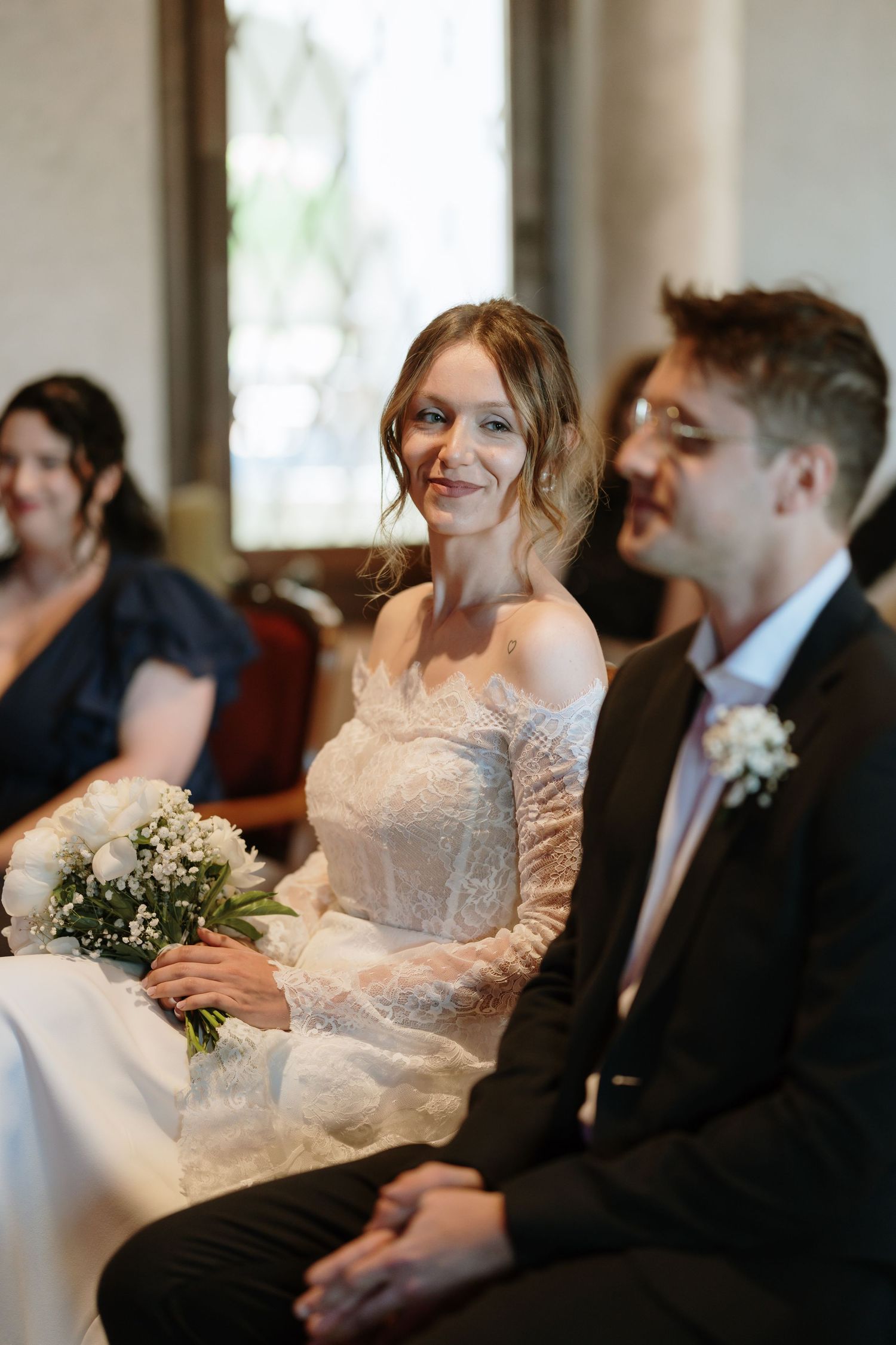 Bride in lace off-shoulder gown smiles while seated with groom during wedding ceremony, holding white bouquet.