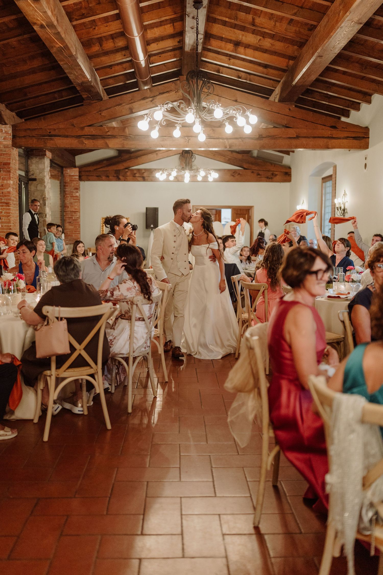Bride and groom share a kiss while entering their rustic wedding reception venue with wooden beamed ceilings and seated guests.