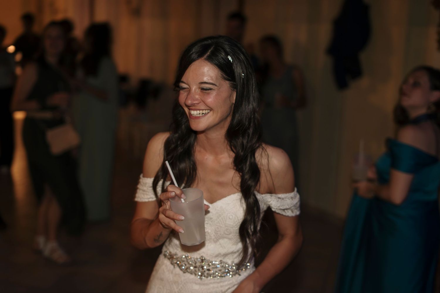 Smiling bride in white off-shoulder gown holding a drink at a wedding reception celebration.