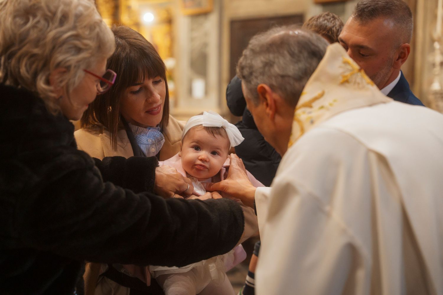 Catholic priest performing baptism ritual on infant baby girl held by family in ornate church.