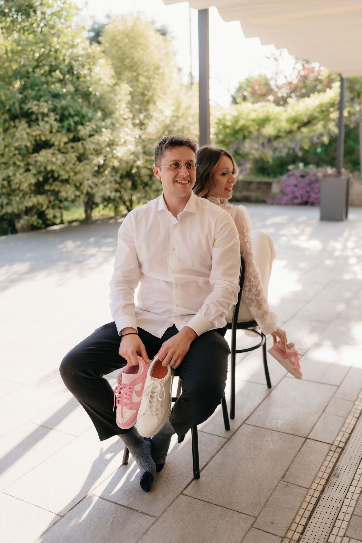 Smiling groom holds pink and white sneakers while bride sits beside him on sunny outdoor terrace.