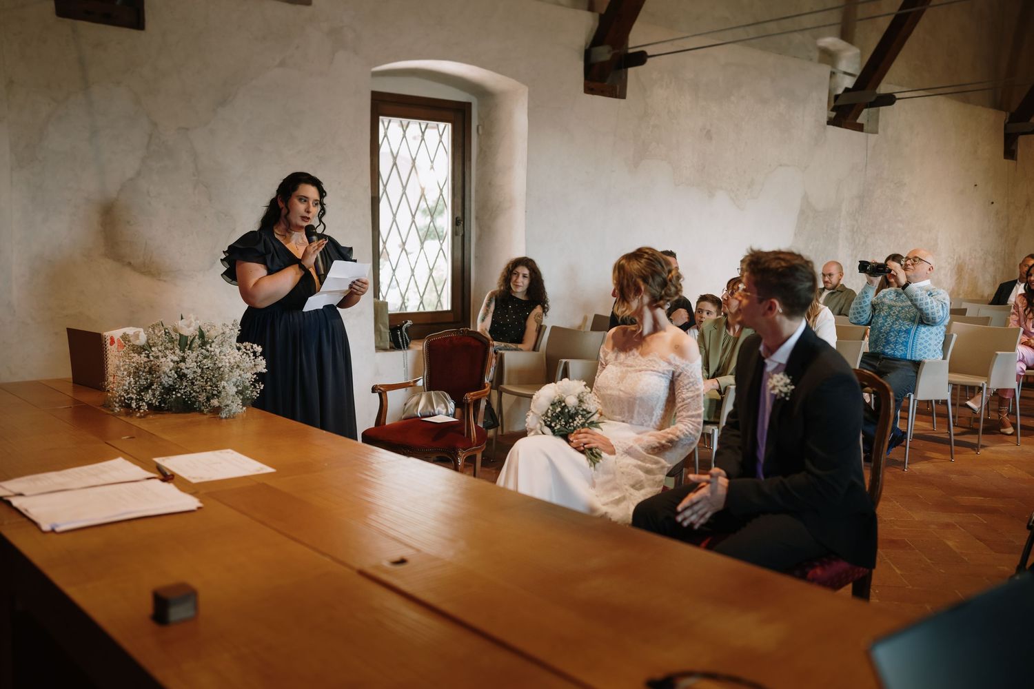 Bride and groom seated at wedding ceremony while officiant reads speech in elegant stone venue.