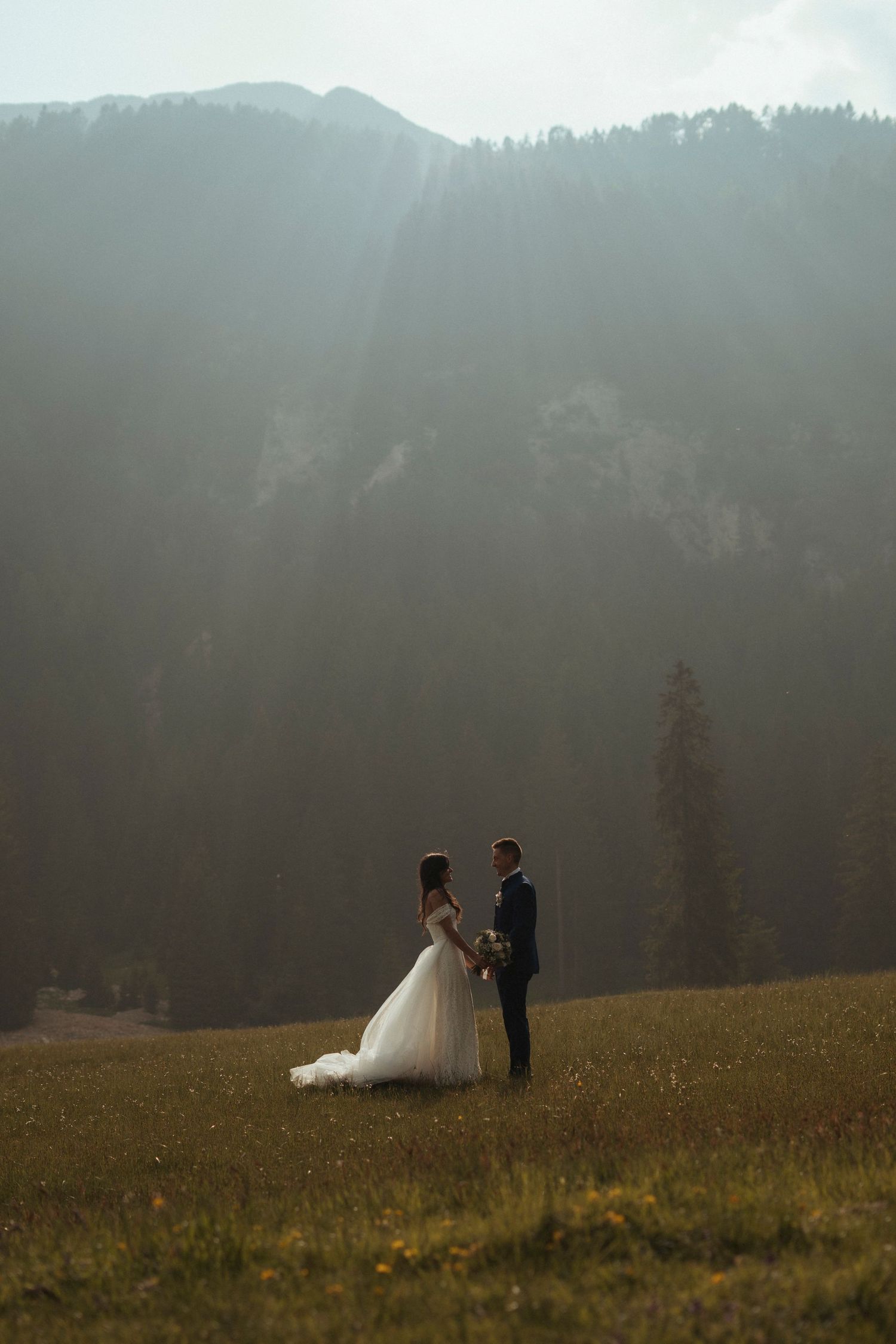 Bride and groom stand in misty mountain meadow with dramatic forested peaks behind them.
