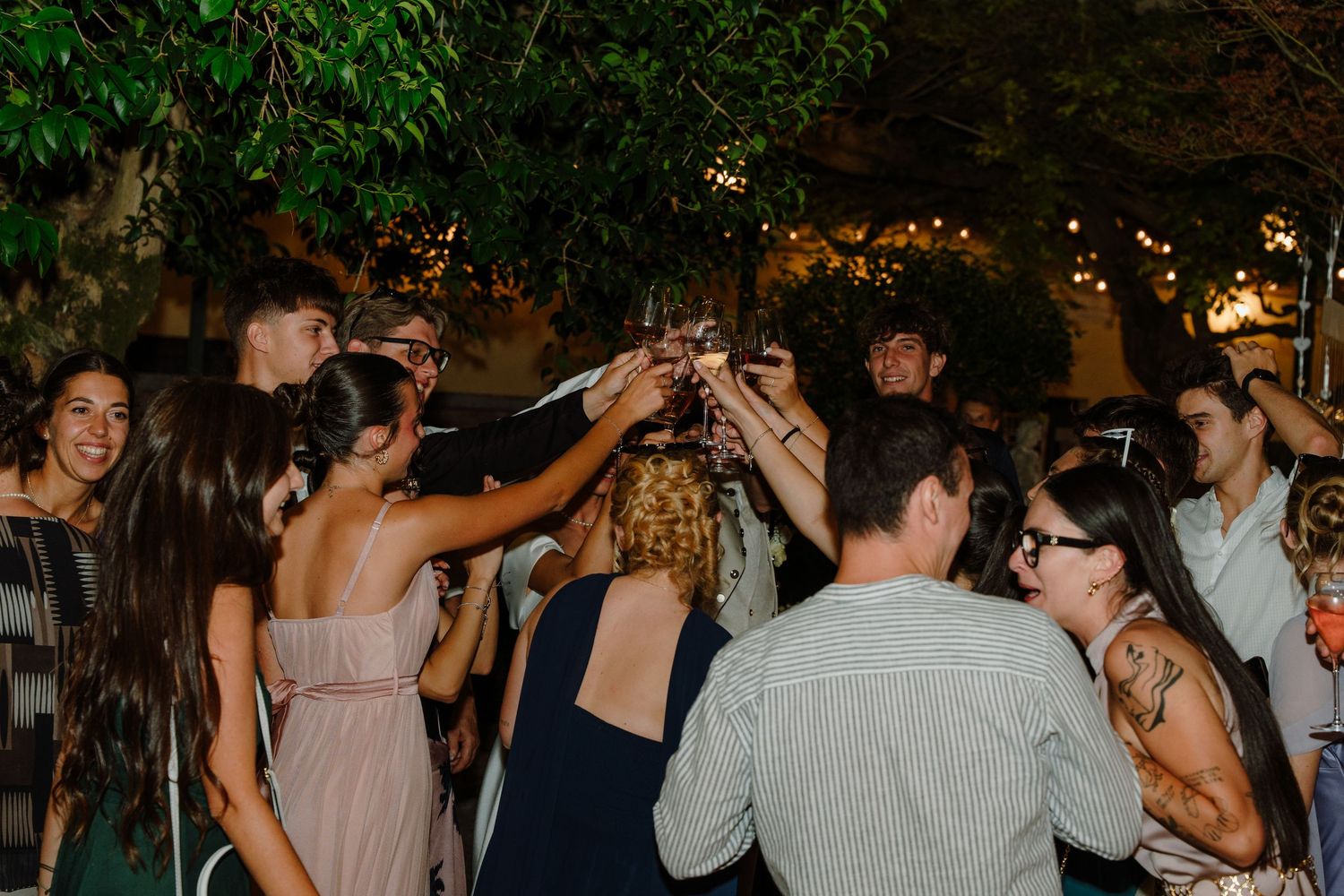 Group of friends raising glasses in a toast at an outdoor evening party surrounded by trees with string lights.