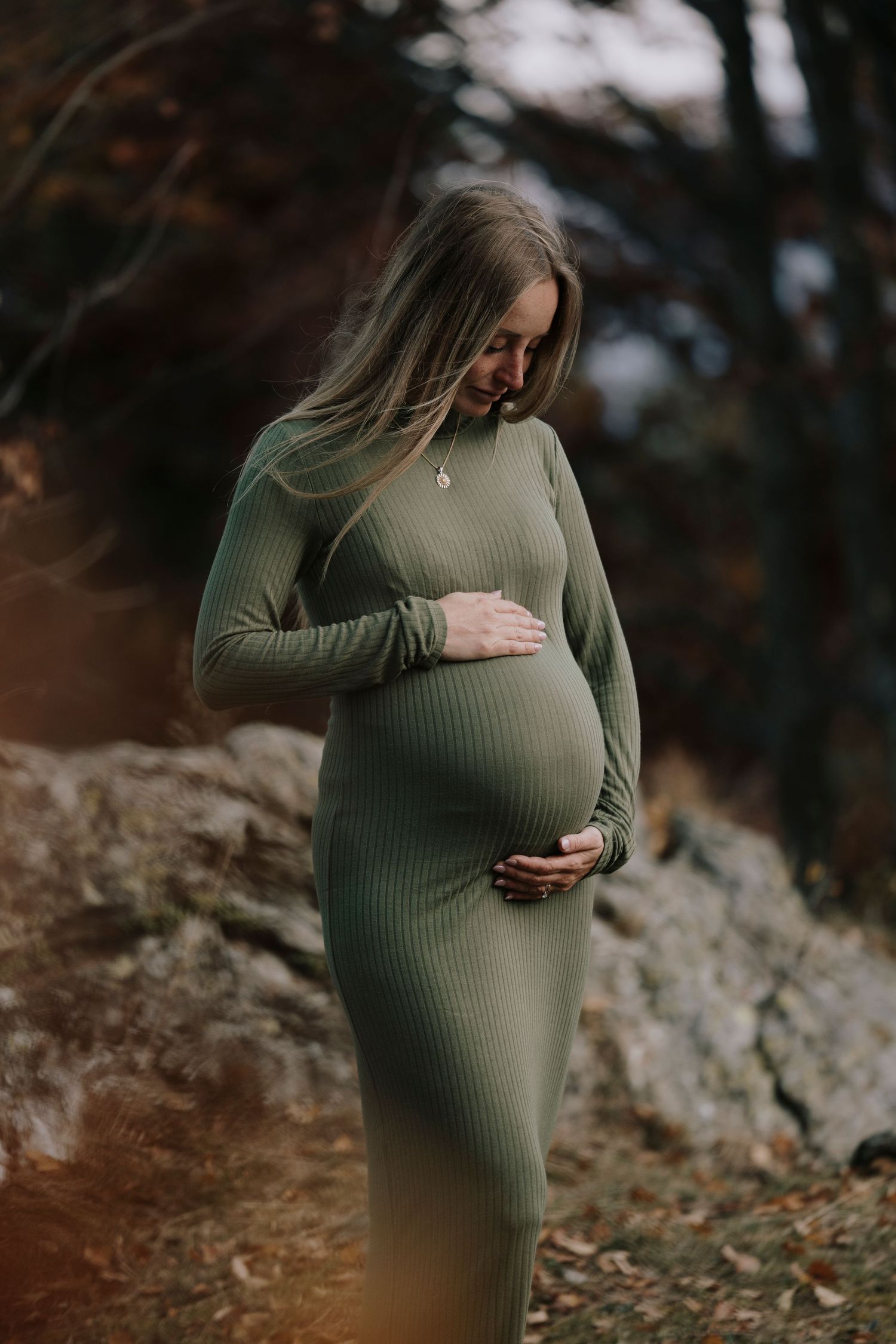 A pregnant woman in a green ribbed dress gently cradles her baby bump in an autumn forest.