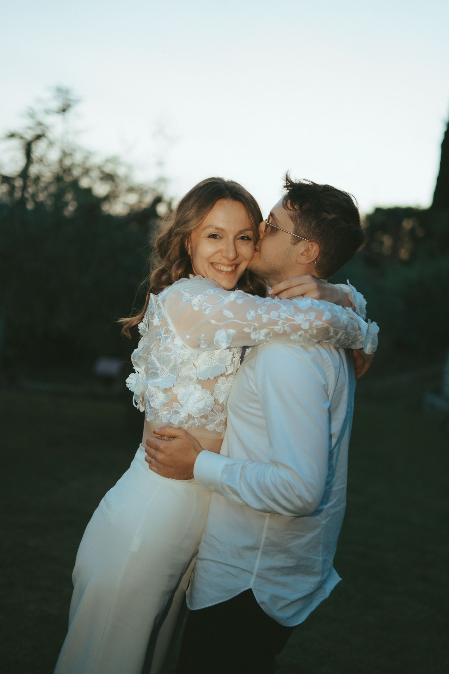 Una sposa sorridente in un abito bianco a motivi floreali, baciata sulla guancia dallo sposo all'aperto, al crepuscolo.