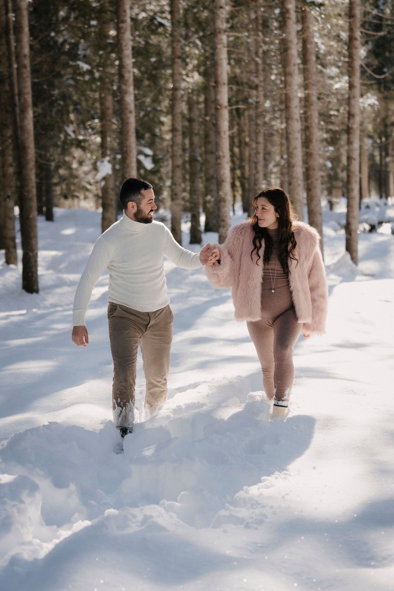 Happy couple holding hands and walking through a snow-covered pine forest on a sunny winter day.